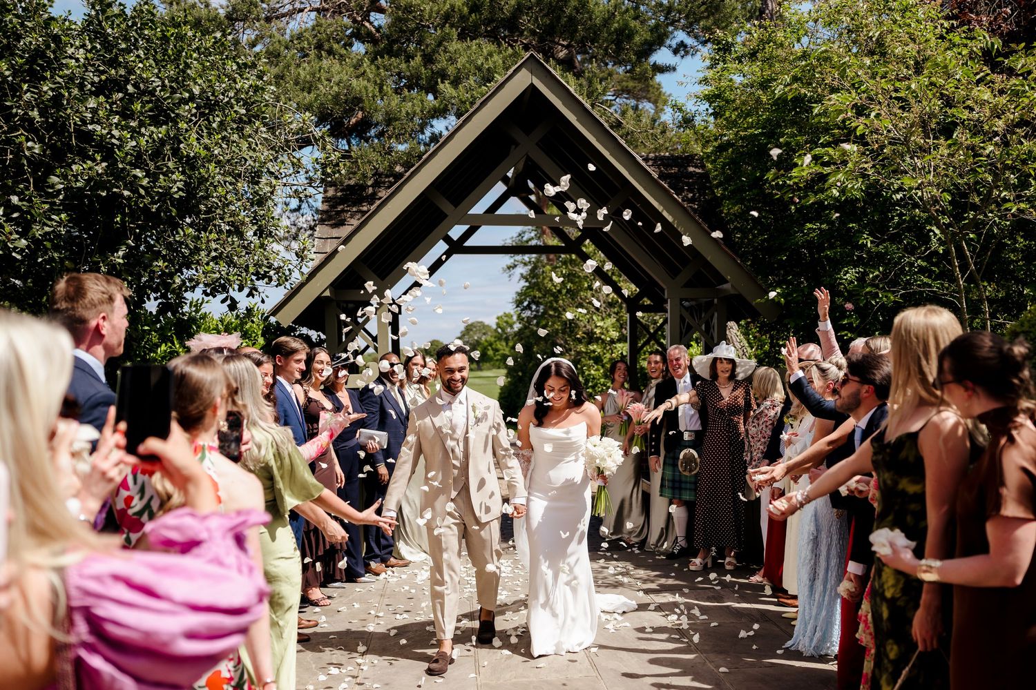 Newlyweds walk through confetti shower under wooden arch surrounded by cheering wedding guests outdoors.