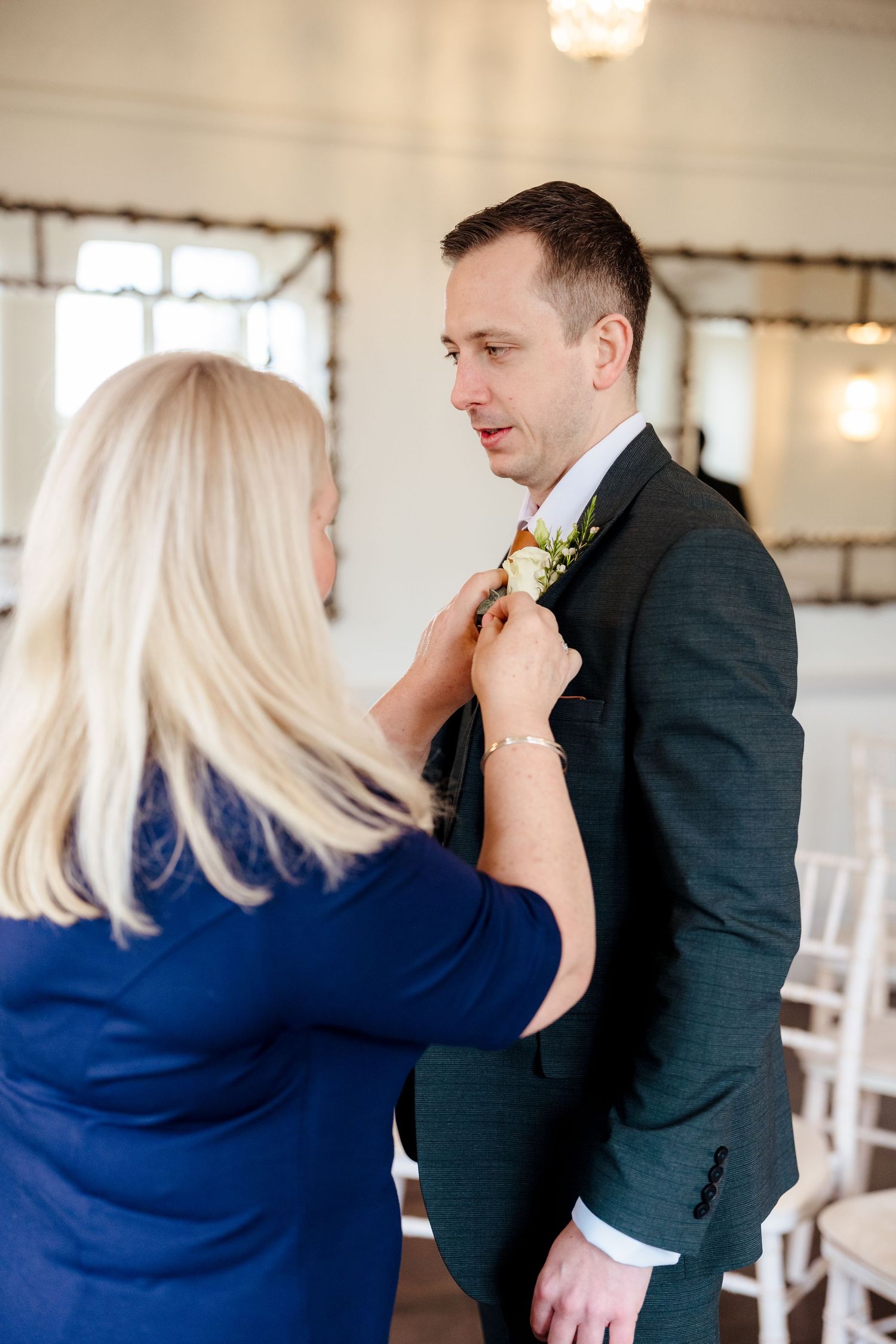 Woman pinning floral boutonniere on groom in dark suit before wedding ceremony.