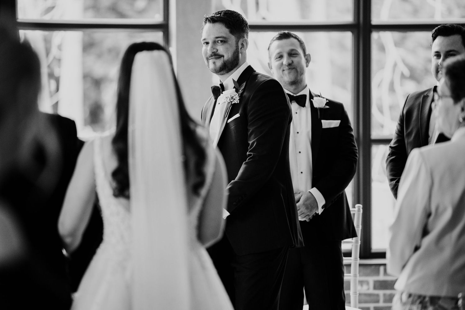 Emotional groom smiling as bride walks down the aisle during an indoor wedding ceremony in black and white photo.