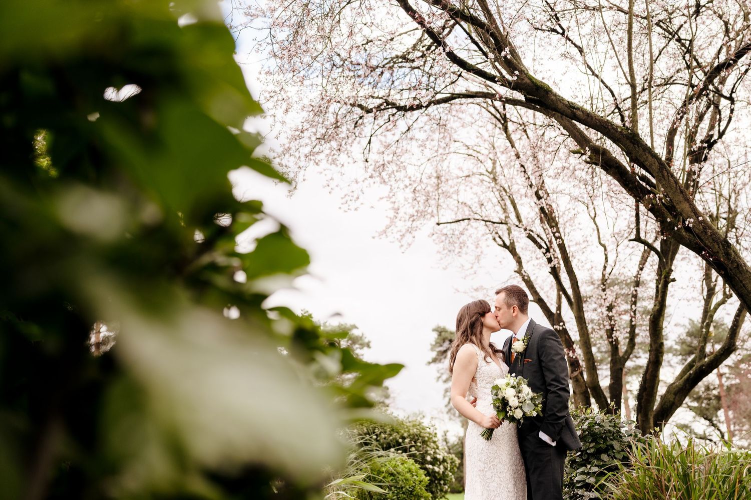 Bride and groom share romantic kiss surrounded by blossoming trees in garden wedding setting.