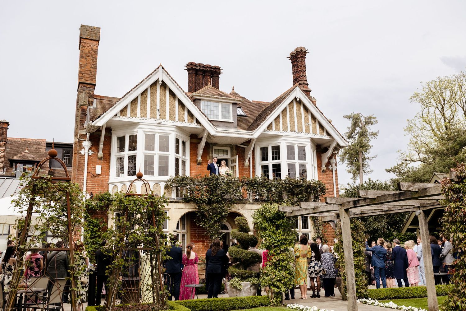 Tudor-style brick wedding venue with guests gathered in a garden adorned with climbing plants and pergolas.