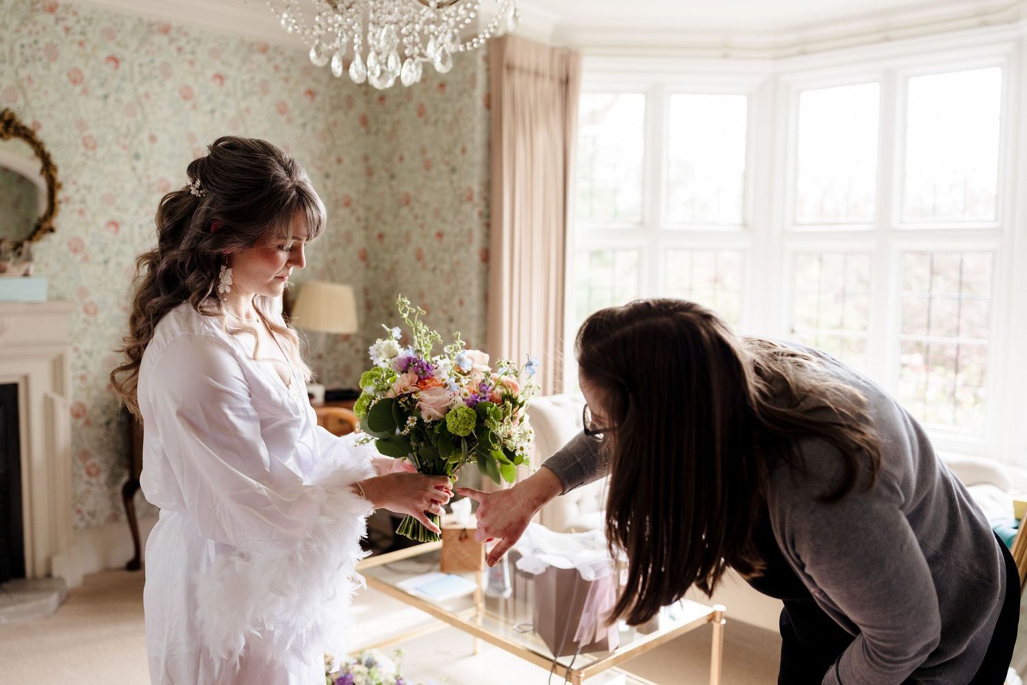 Bride in white robe receiving floral bouquet from assistant in elegant floral-wallpapered room with chandelier.