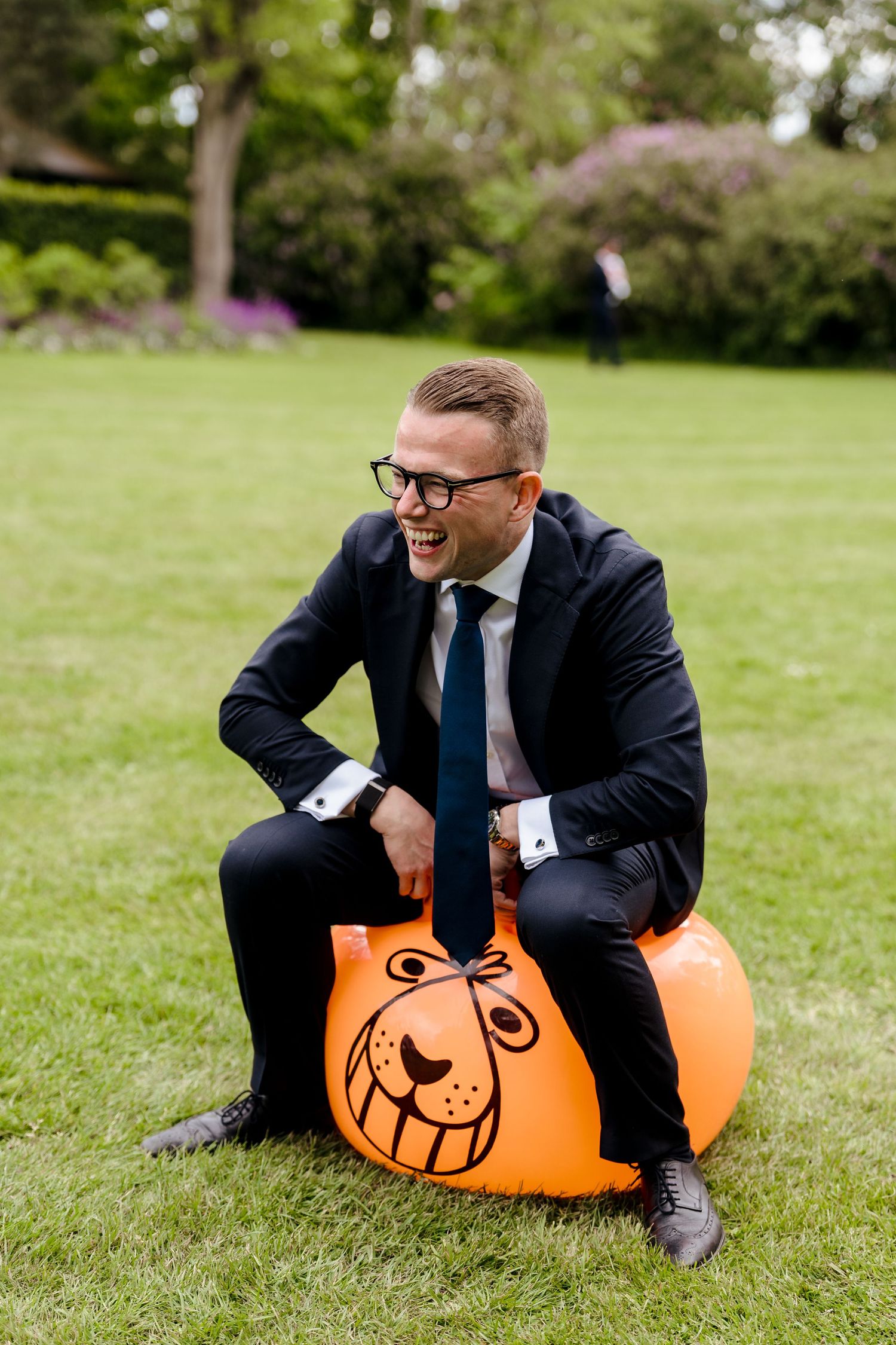 Laughing groomsman in navy suit sits on orange inflatable bouncy hopper toy on green lawn at wedding reception.