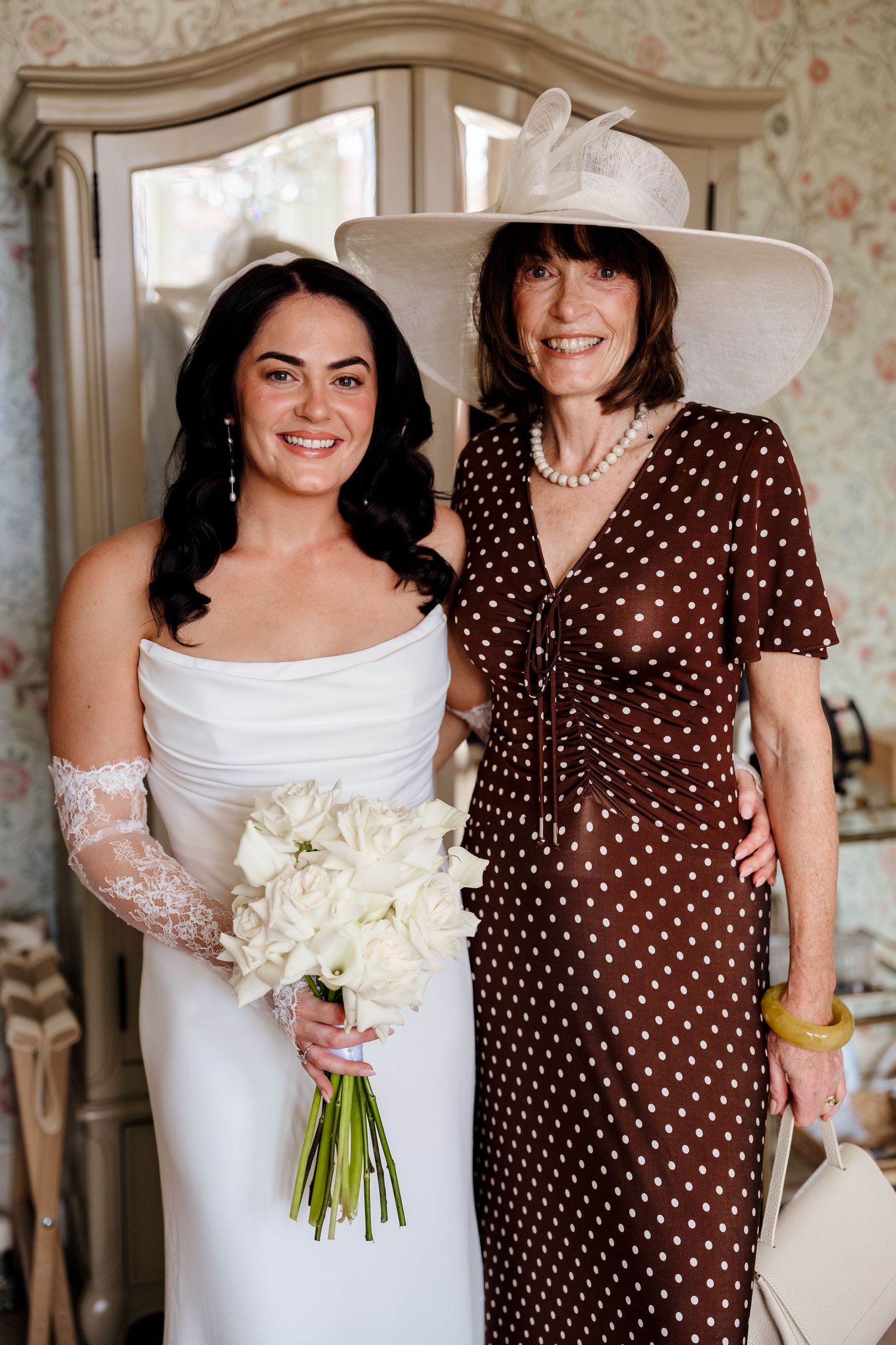 Bride in white strapless gown holding white bouquet poses with older woman in brown polka dot dress and wide-brim hat.