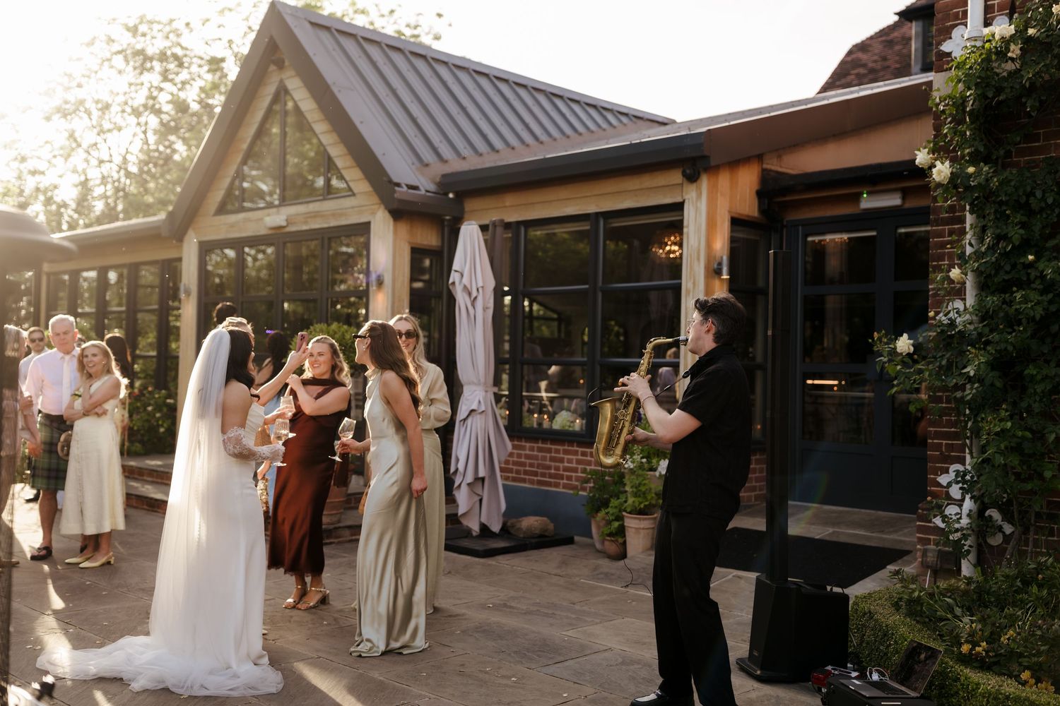 Bride and guests enjoying live saxophone performance on sunny outdoor patio at wedding venue.