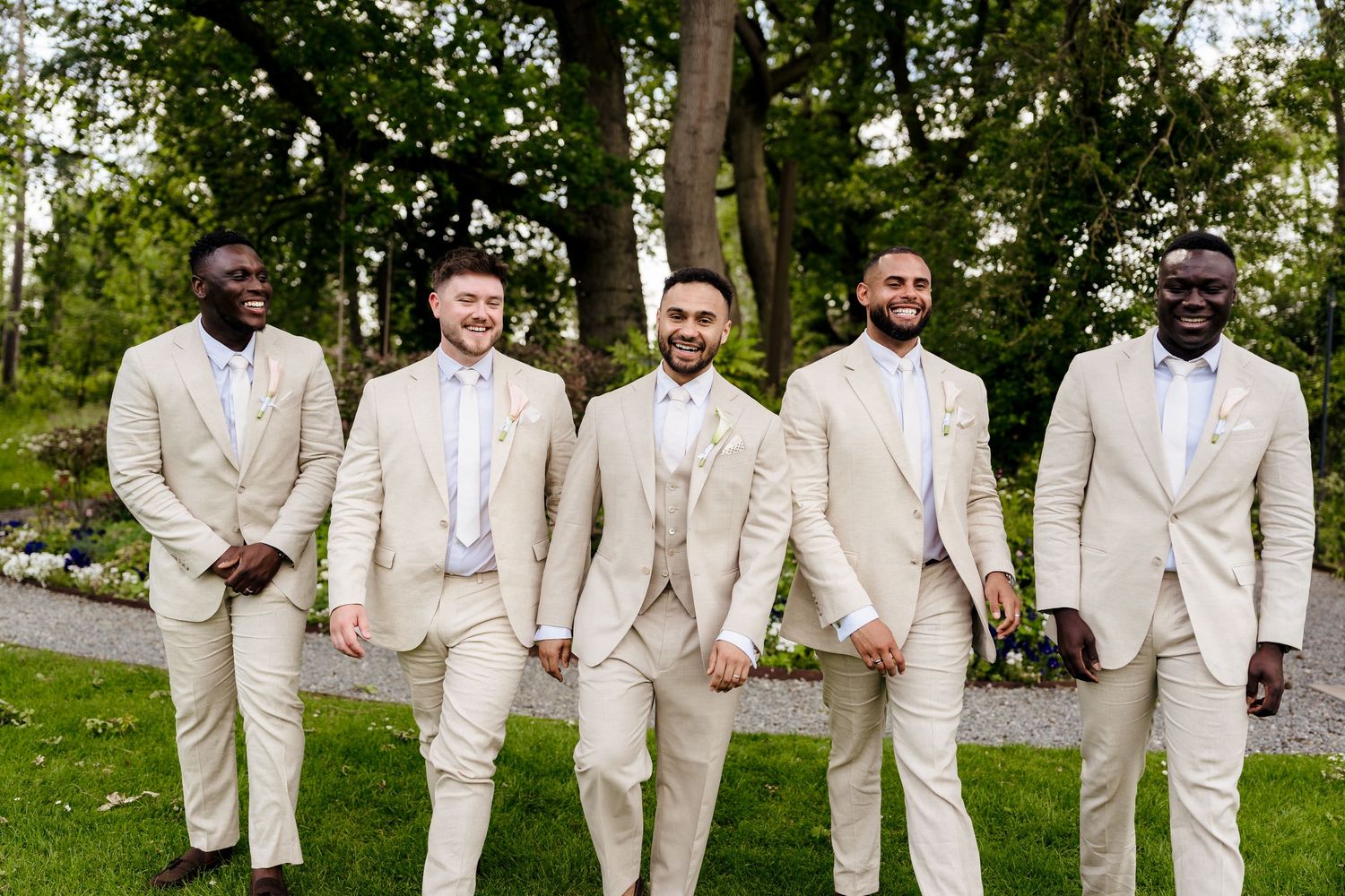 Groom and four groomsmen in matching beige suits smiling together outdoors at wedding celebration.