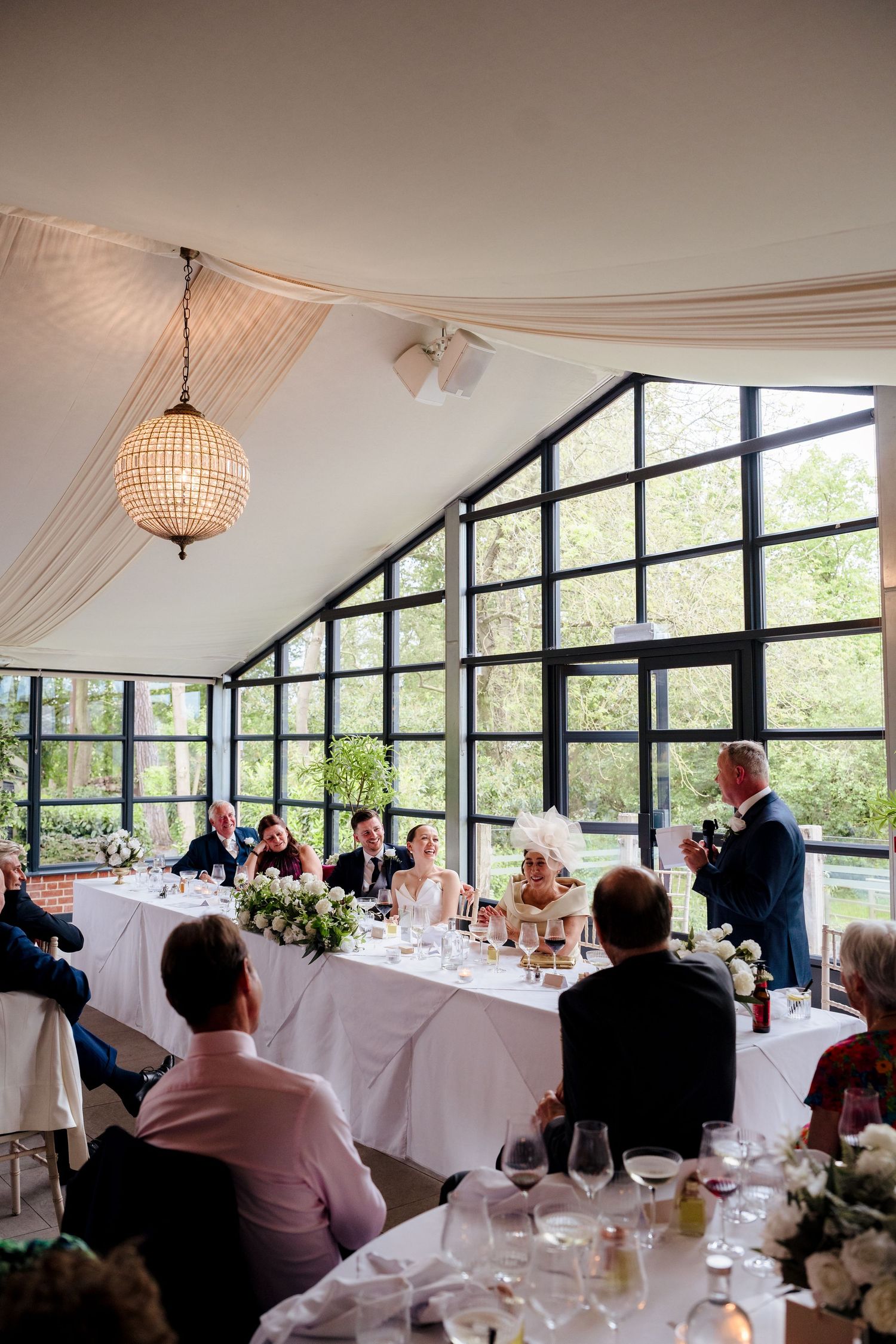 Guests enjoy a wedding speech in an elegant glass-walled reception venue with floral centerpieces.