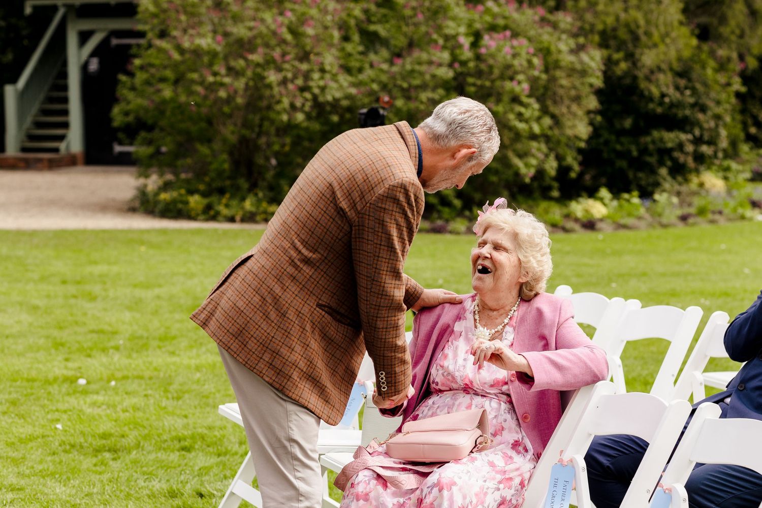 Elderly couple sharing a joyful laugh outdoors at a garden event, woman in pink seated on white chair.