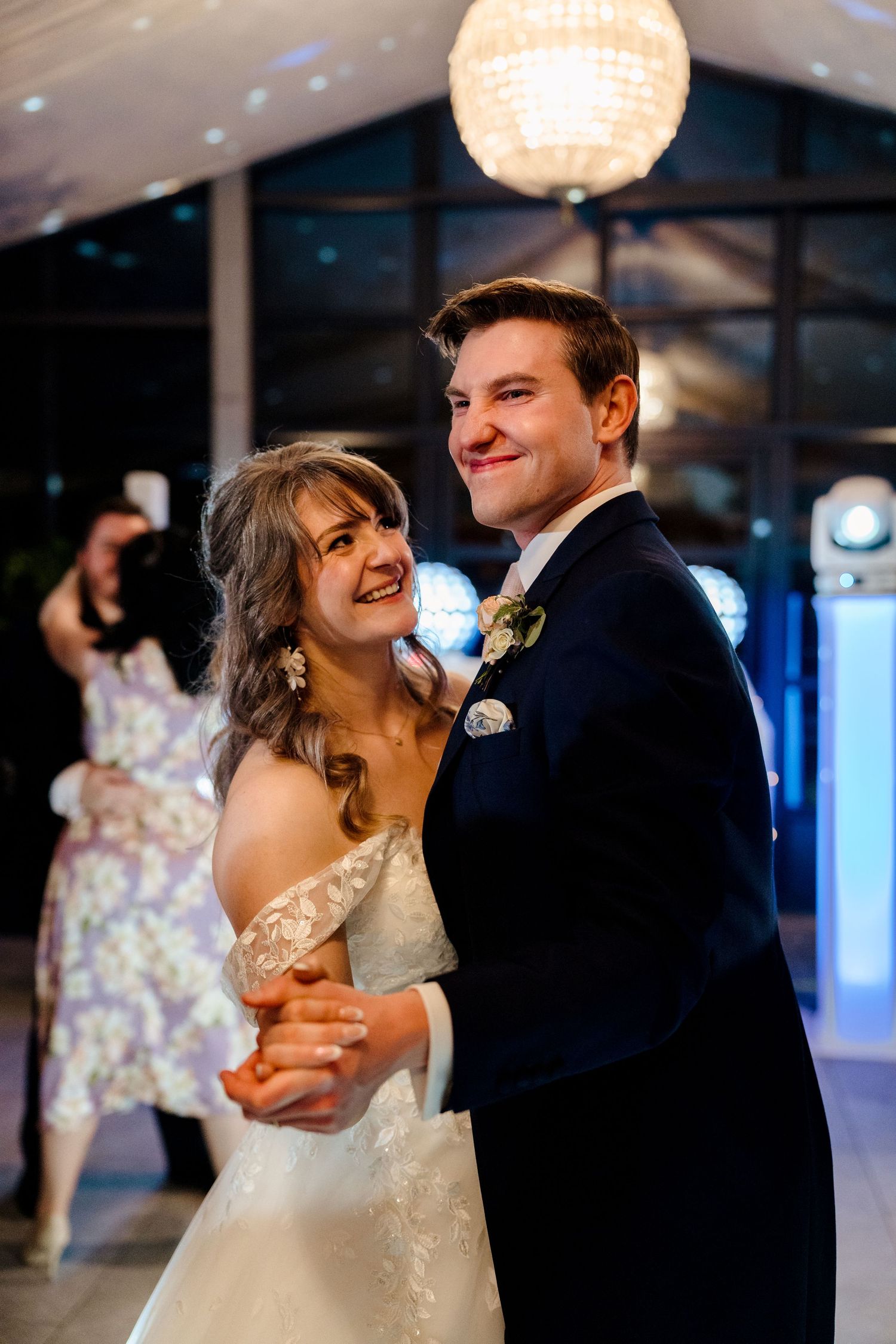 Bride and groom share their first dance, smiling and holding hands under a glowing chandelier at their wedding reception.