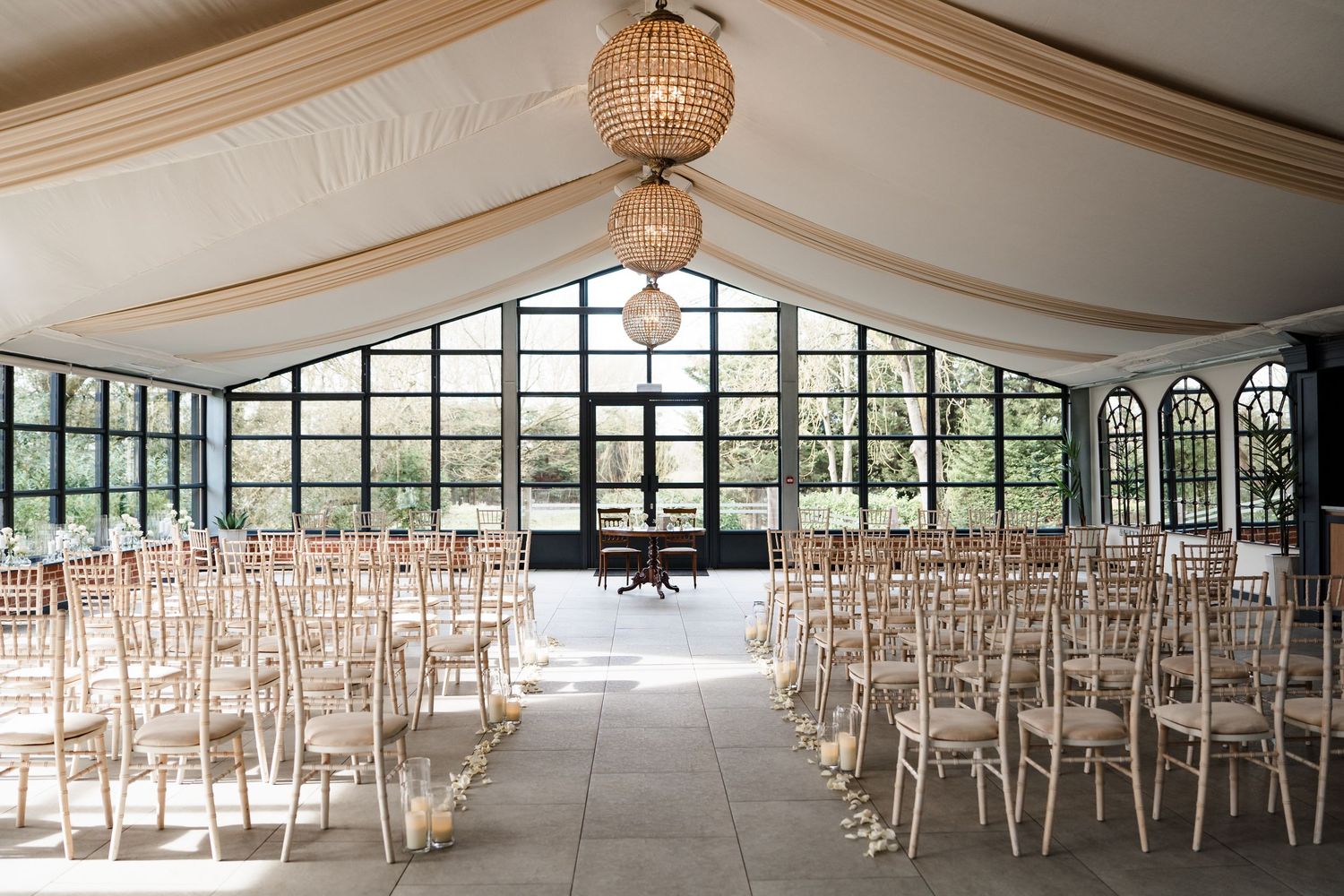 Elegant wedding ceremony venue with rows of chiavari chairs, draped ceilings, chandelier lighting, and large greenhouse-style windows.
