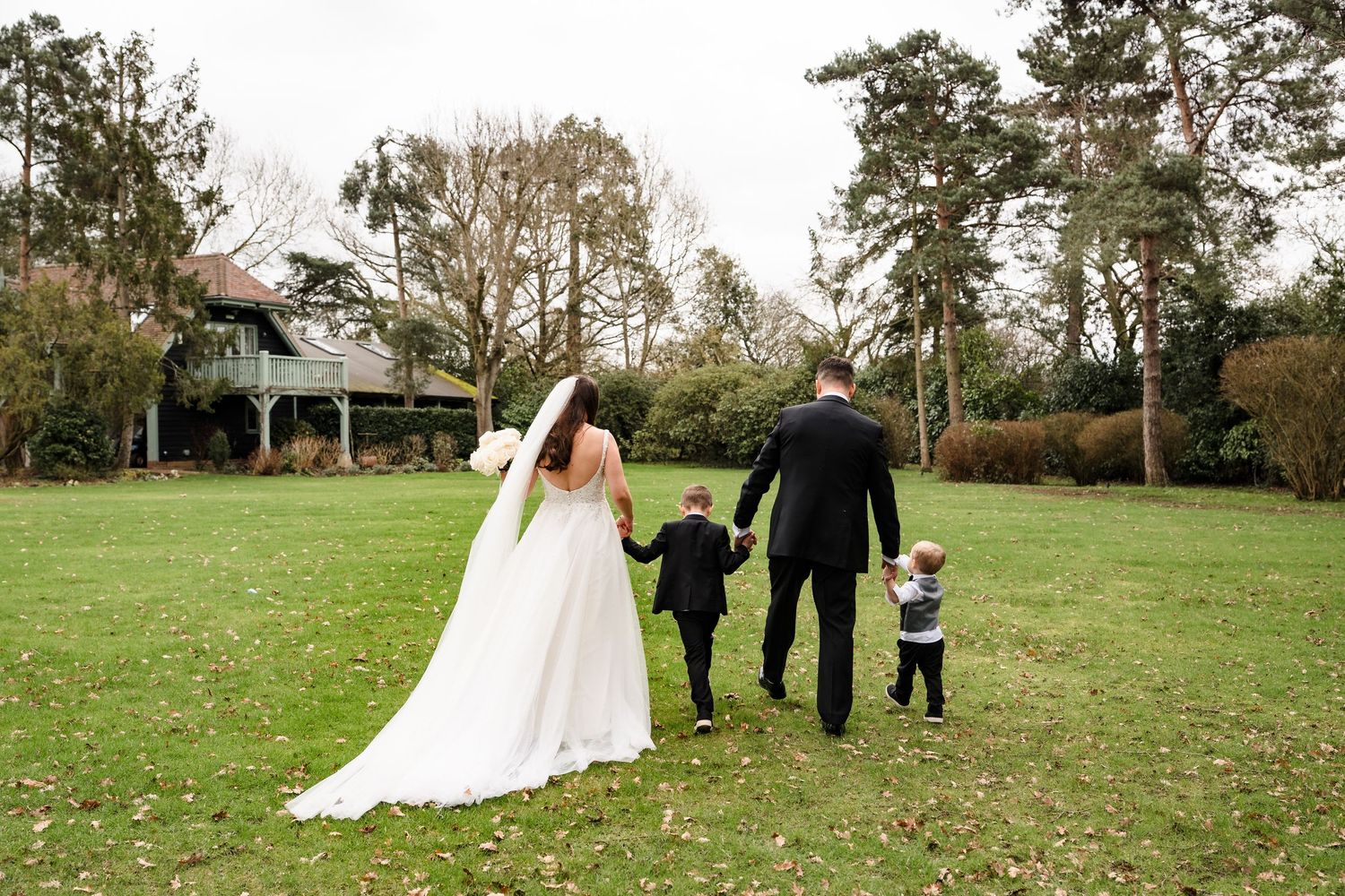 Bride and groom walk hand in hand with two young children across a lush green lawn.