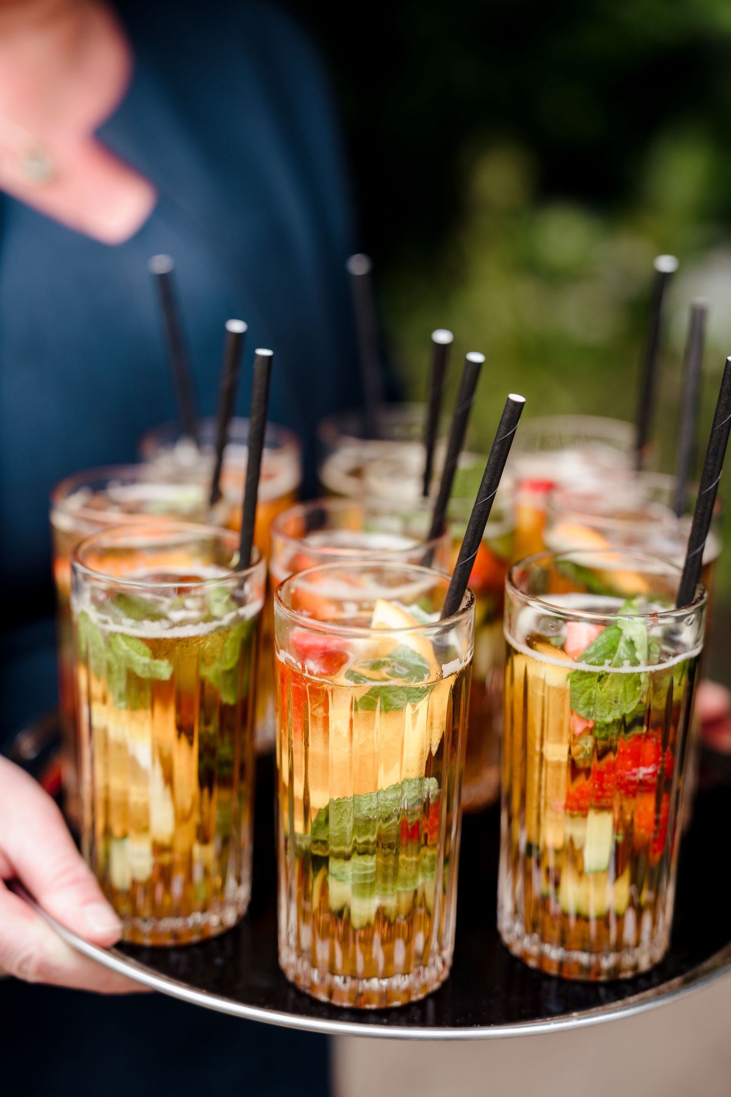 Elegant tray of colorful cocktails garnished with fresh fruit, mint, and black straws at wedding reception.