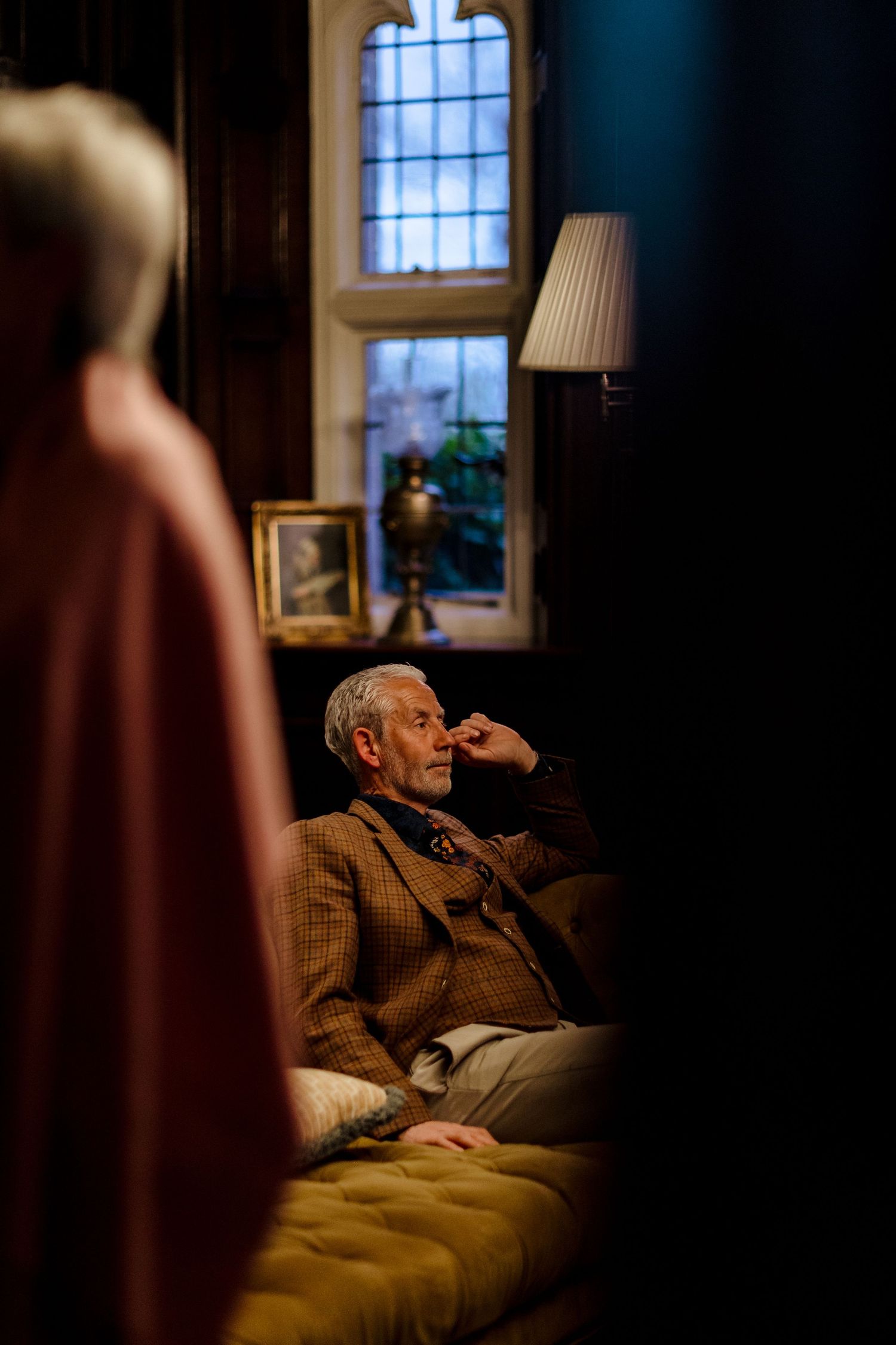 Elderly man in tweed jacket sitting pensively on tufted sofa in dimly lit vintage room.