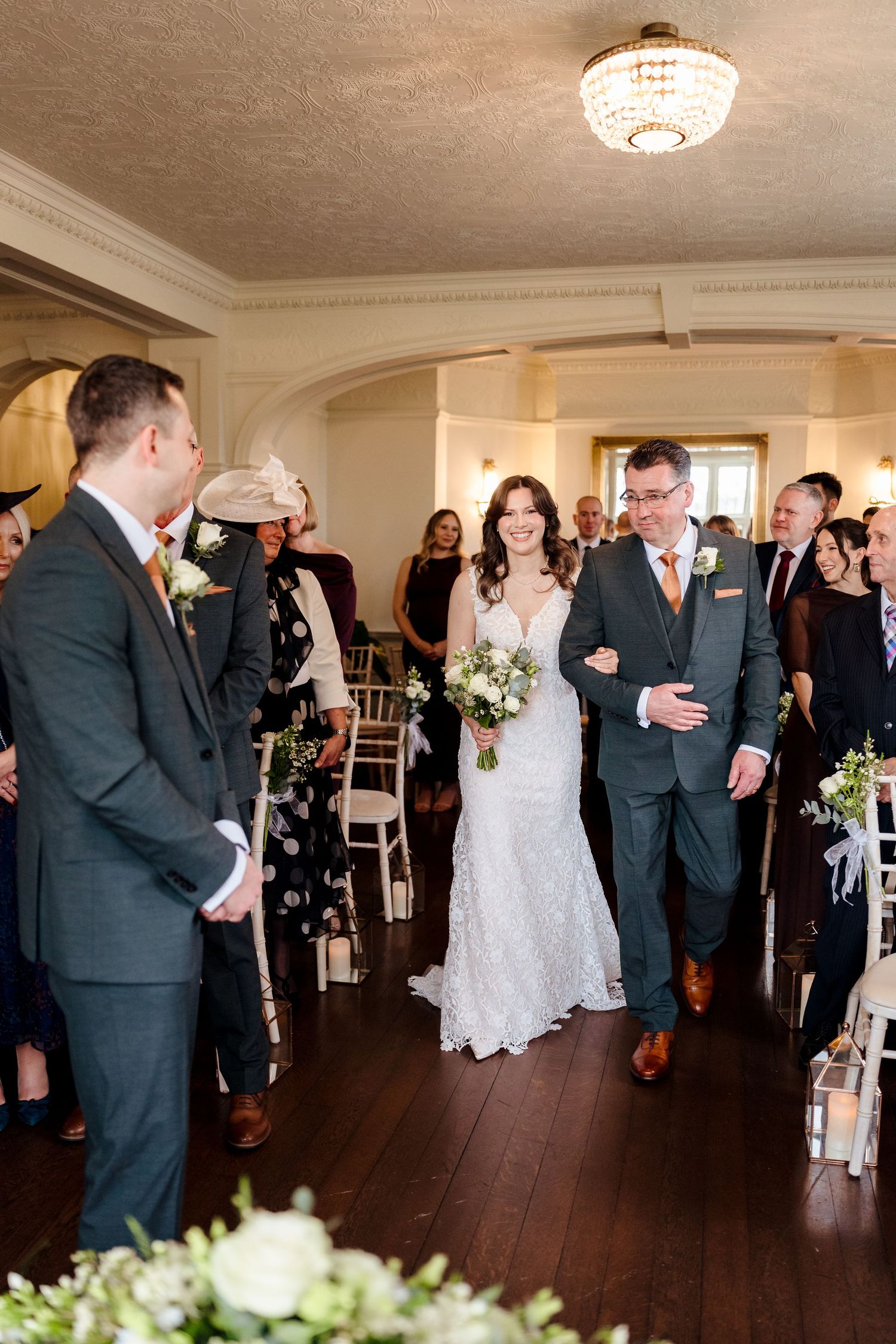 Bride walks down the aisle in lace gown escorted by man in grey suit.