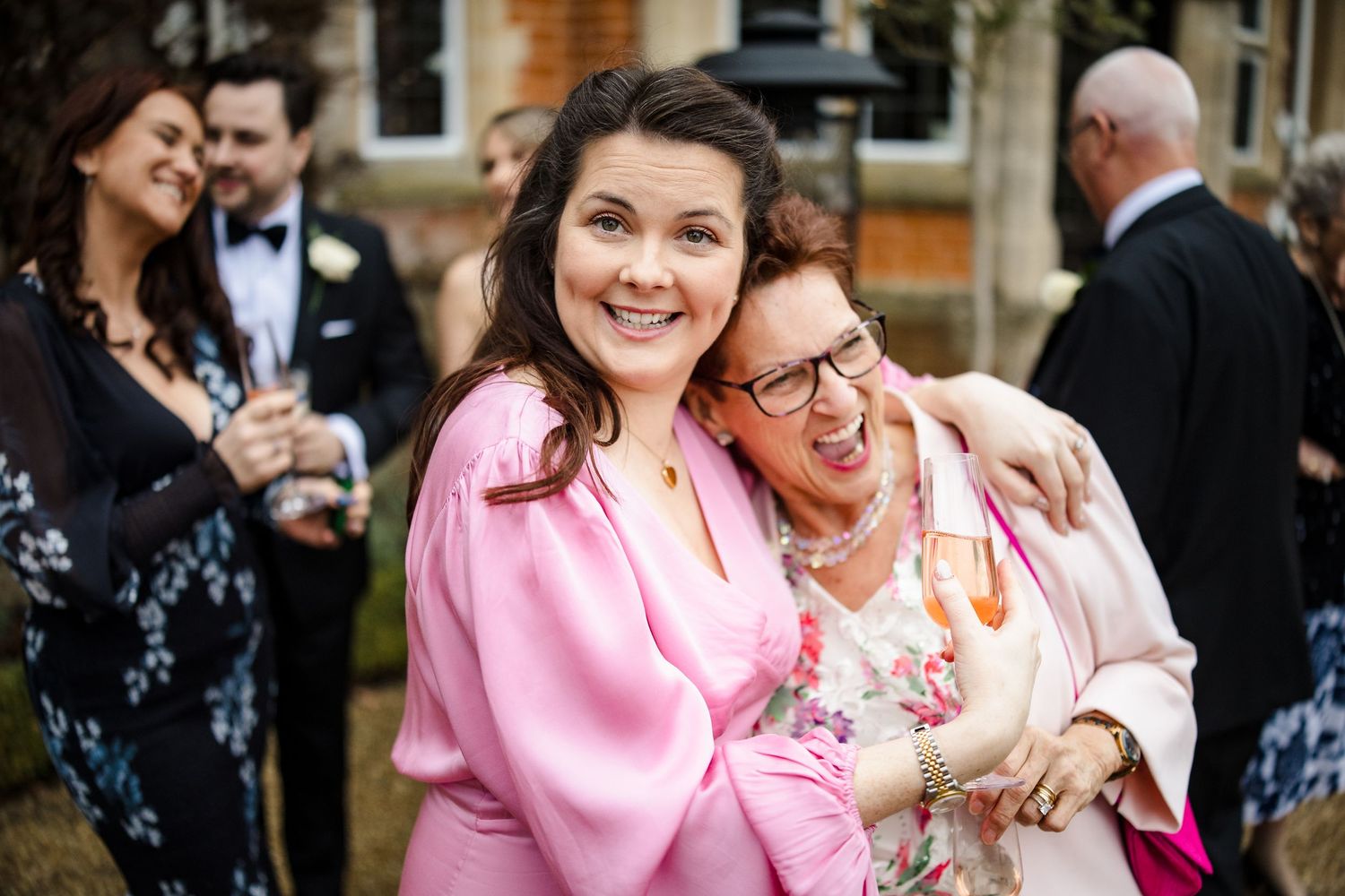 Two women smiling and embracing at an outdoor wedding celebration with guests in the background.