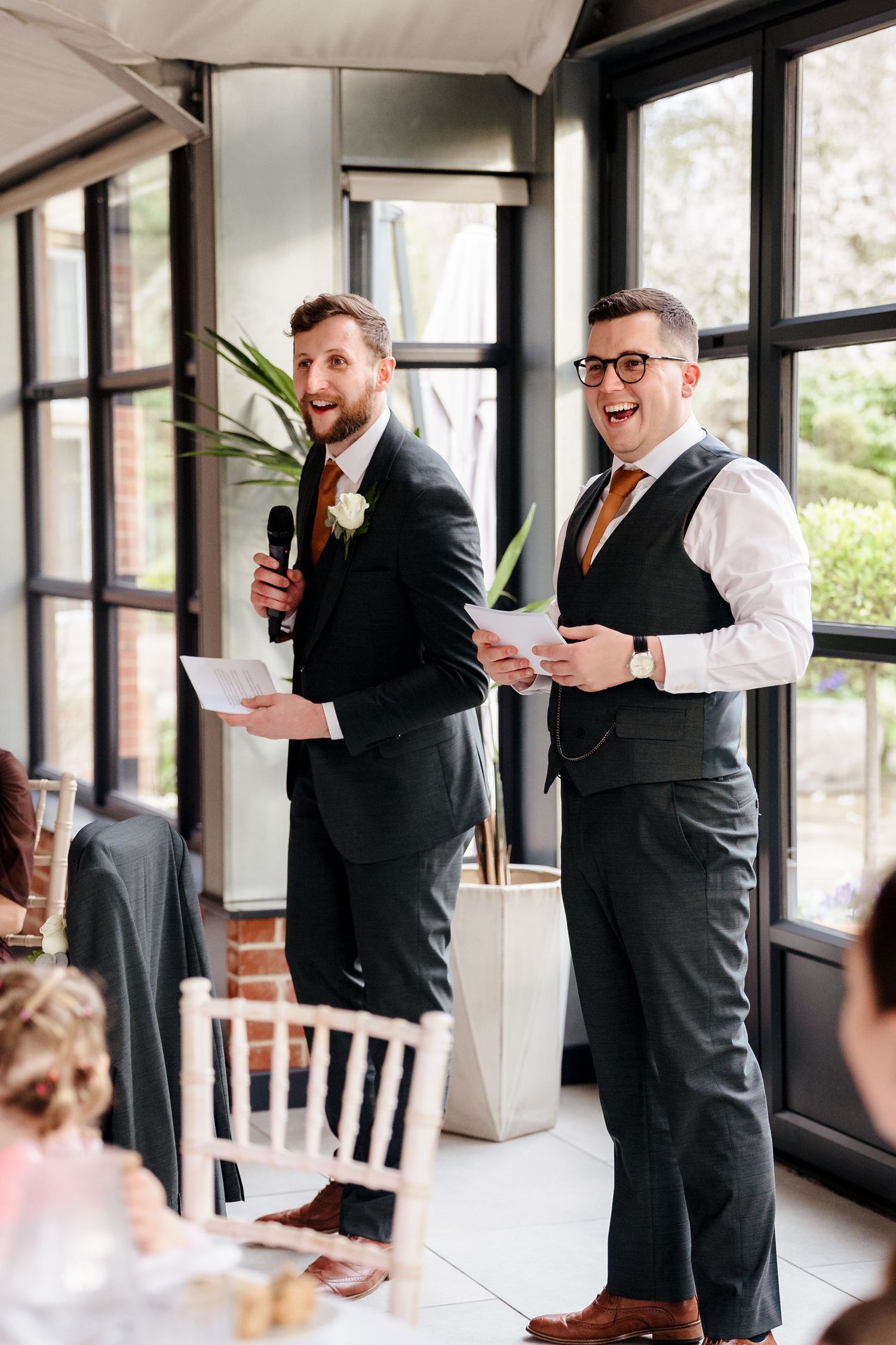 Two best men laugh while delivering a joint wedding speech holding microphone and notes.