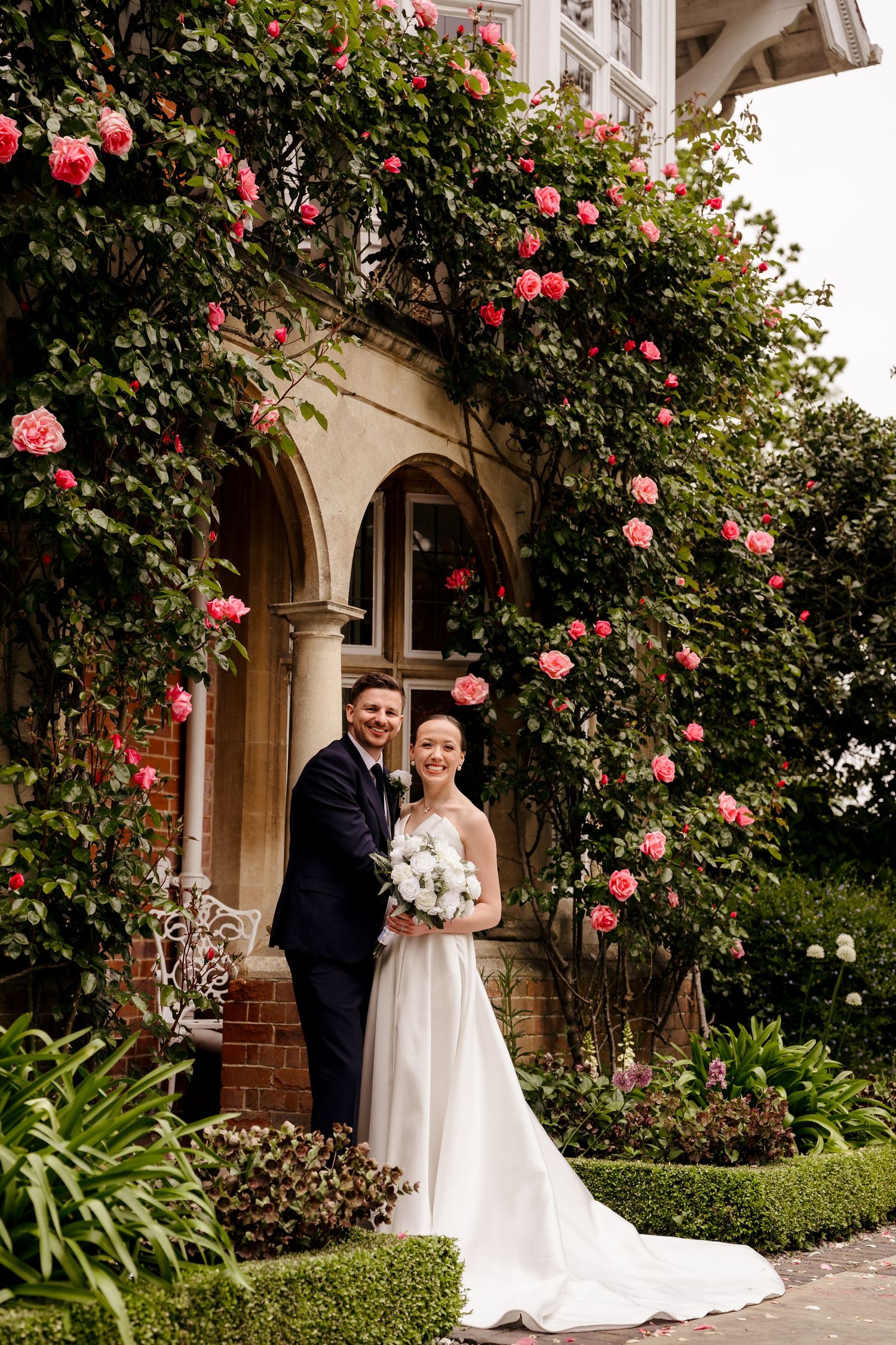 Bride and groom pose beneath climbing pink roses on historic brick building exterior.