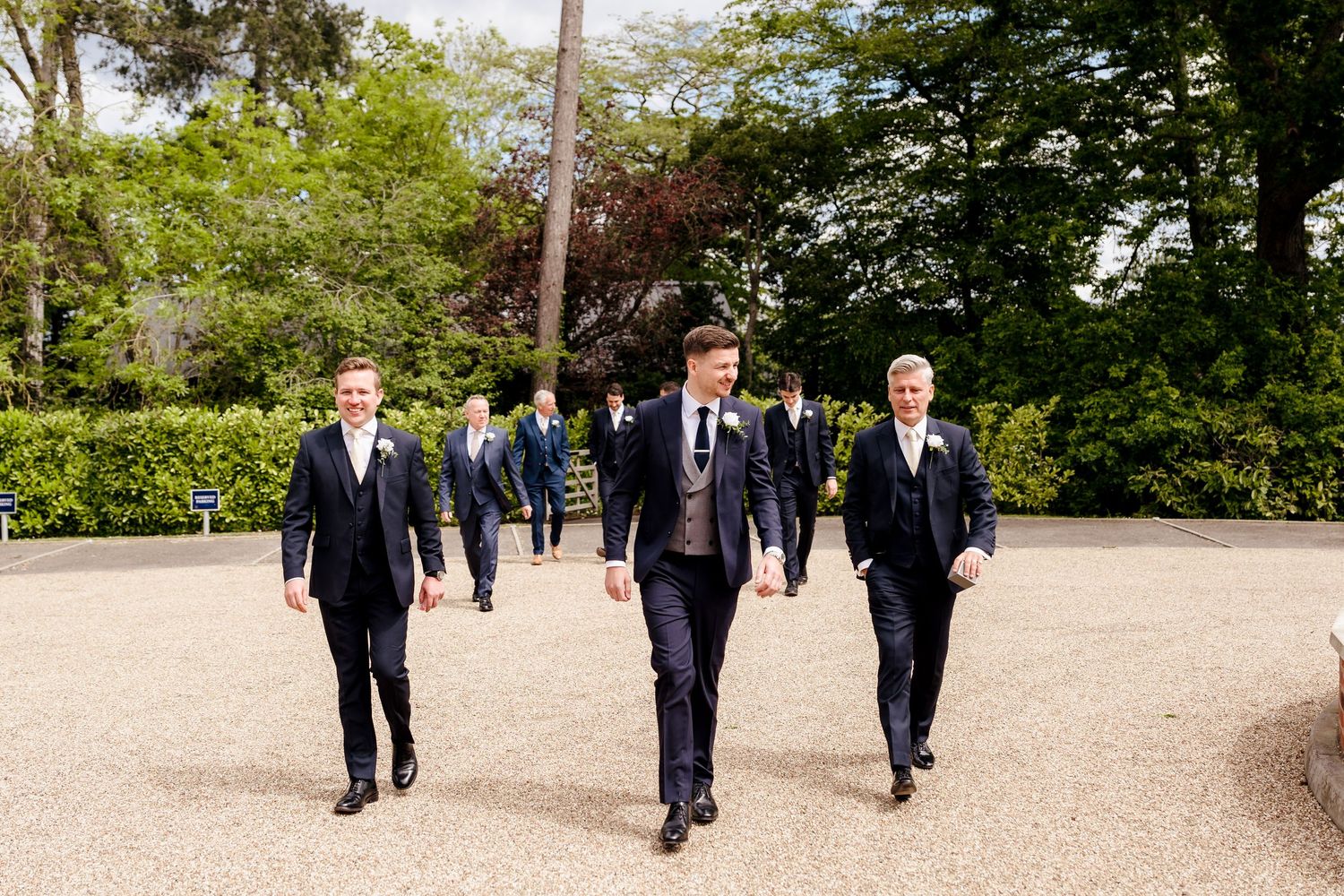 Groomsmen in navy suits walking confidently along a gravel path surrounded by lush green trees at a wedding.
