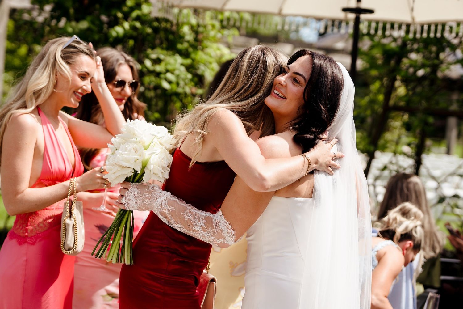 Bride in white gown hugging bridesmaid in red dress while guests in pink dresses celebrate outdoors.