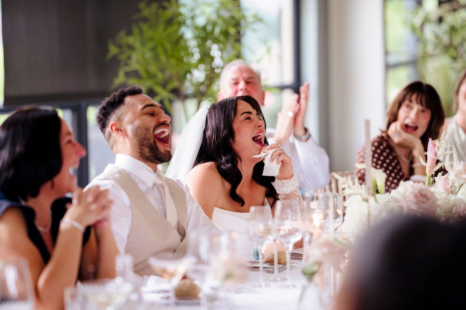 Bride and groom laughing joyfully with guests at wedding reception dinner table with glassware.