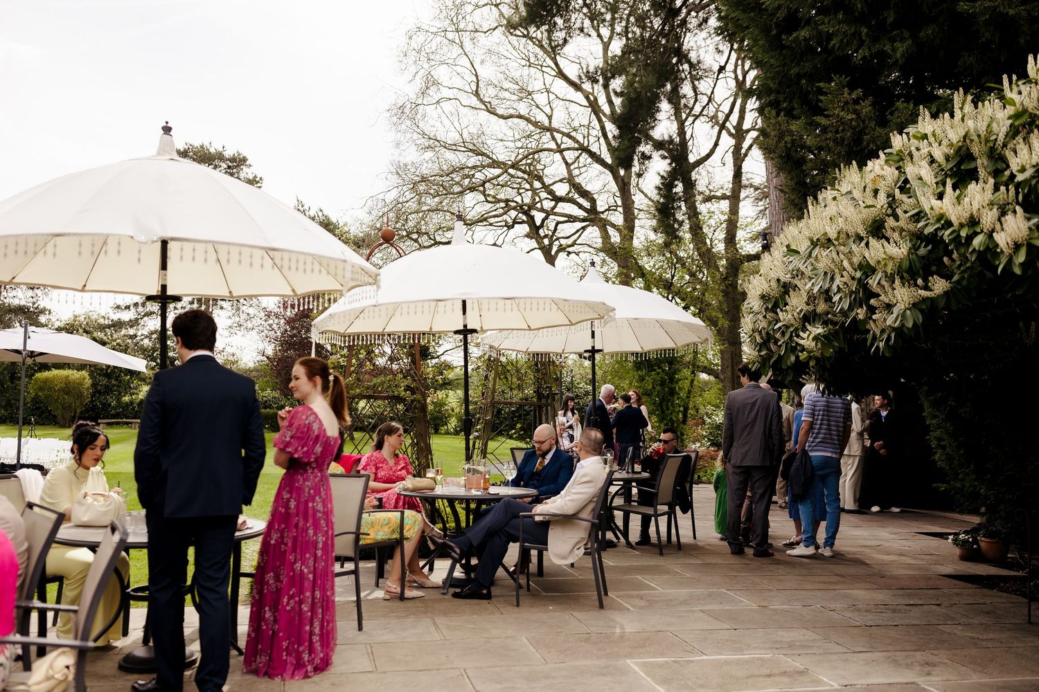 Wedding guests mingling on outdoor patio with white parasols and lush garden backdrop.