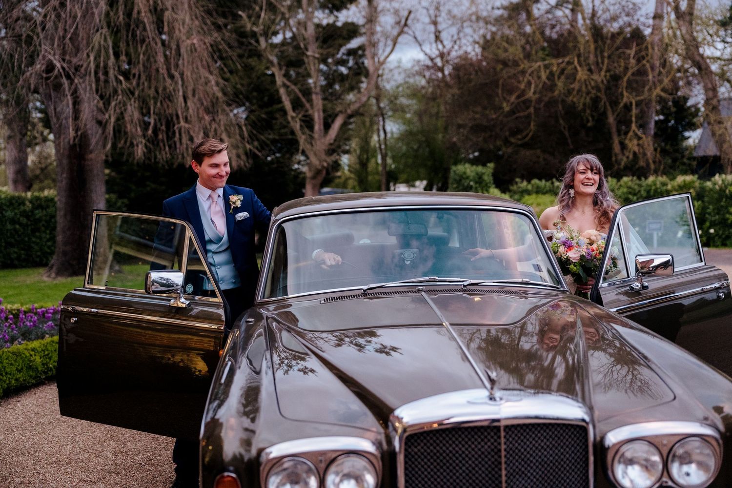 Bride and groom smiling beside a classic black Bentley luxury car in a scenic outdoor wedding venue.