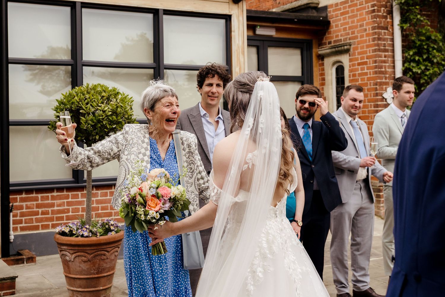 Excited elderly woman raises champagne glass to celebrate with bride in white gown outdoors.