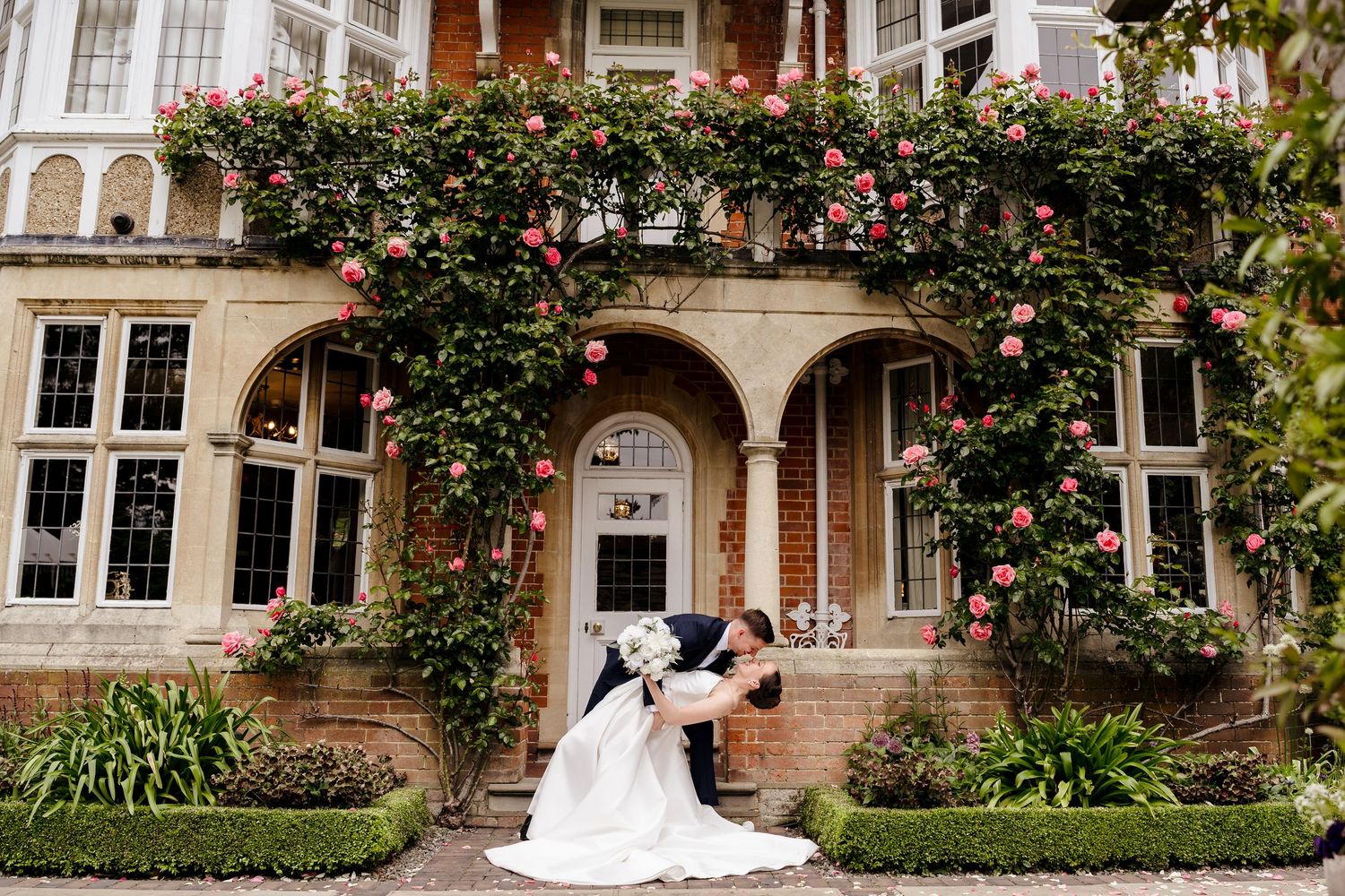 Bride and groom romantic dip kiss in front of rose-covered brick manor house wedding venue.