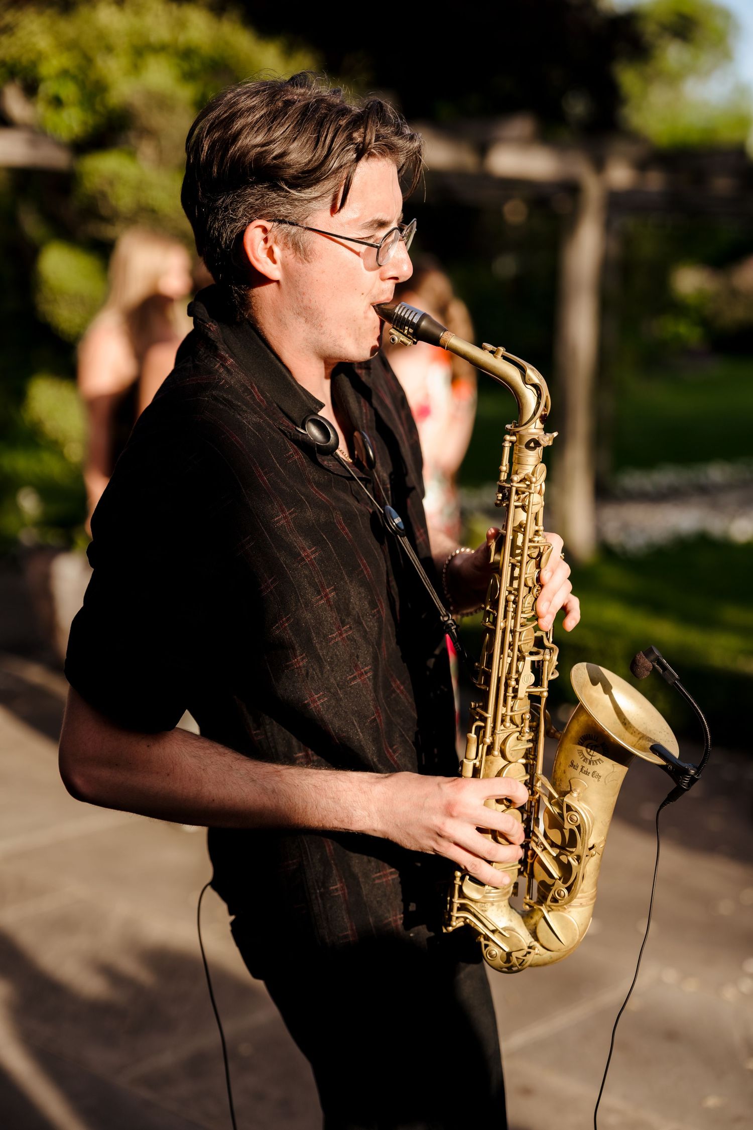 Male saxophonist in black shirt performs outdoors at a wedding reception in a garden setting.