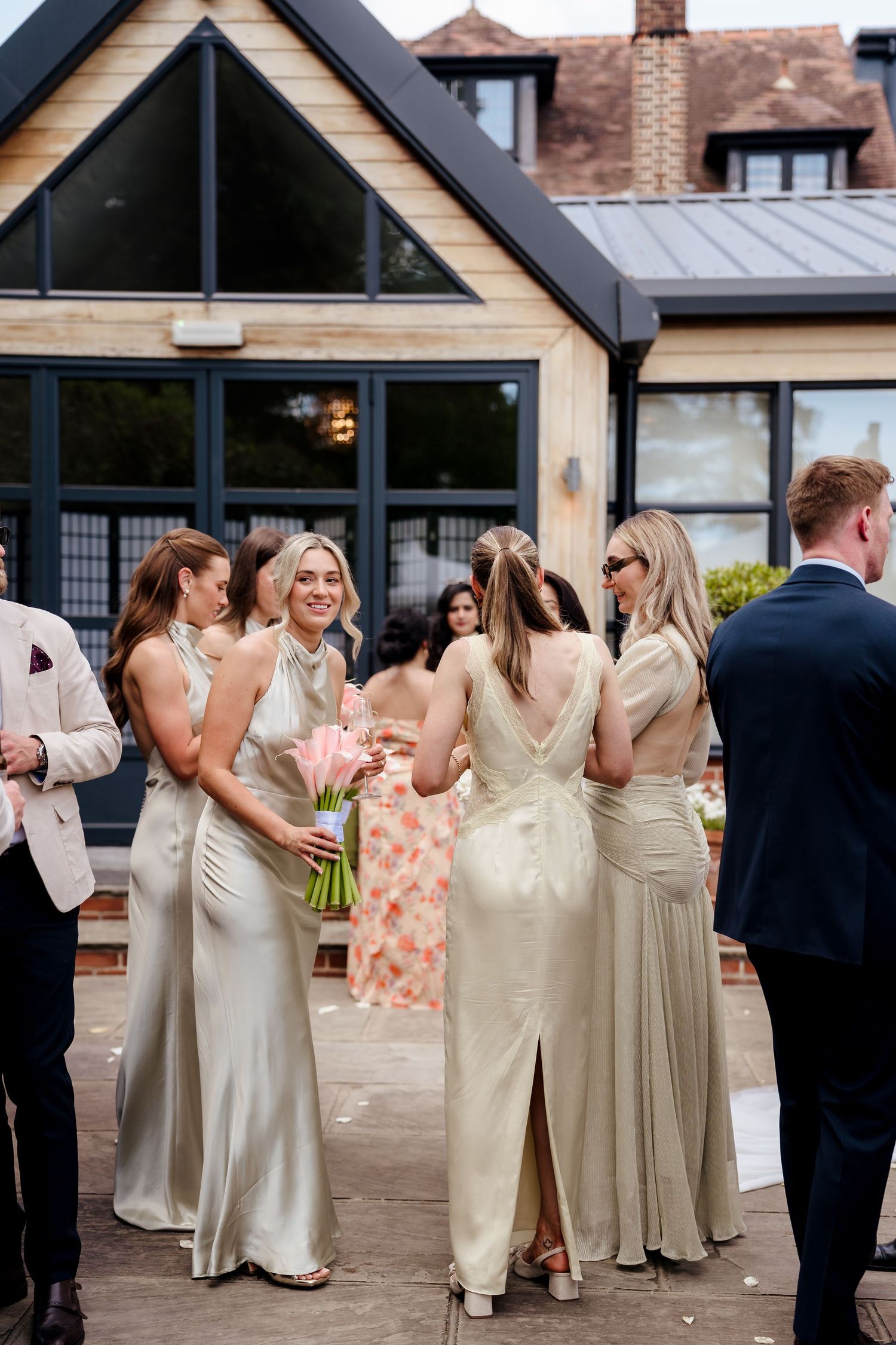Bridesmaids in elegant champagne satin gowns gather outside a modern wedding venue holding pink bouquets.