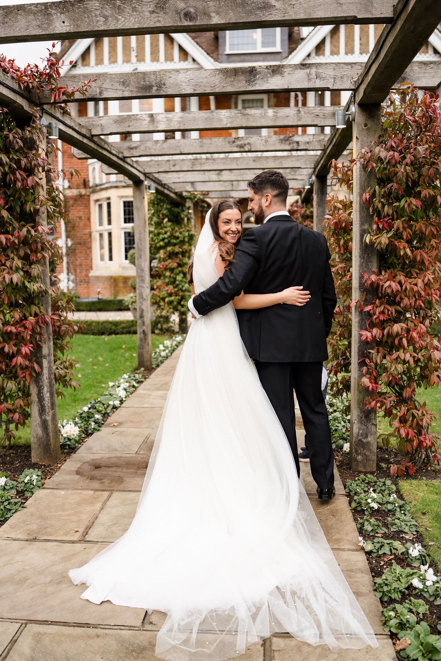 A bride in a white gown and groom in a black suit embrace under a wooden pergola in a lush garden setting.