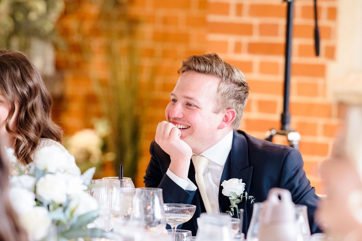 Smiling groom in a navy suit with a white boutonniere laughing at the wedding reception table.