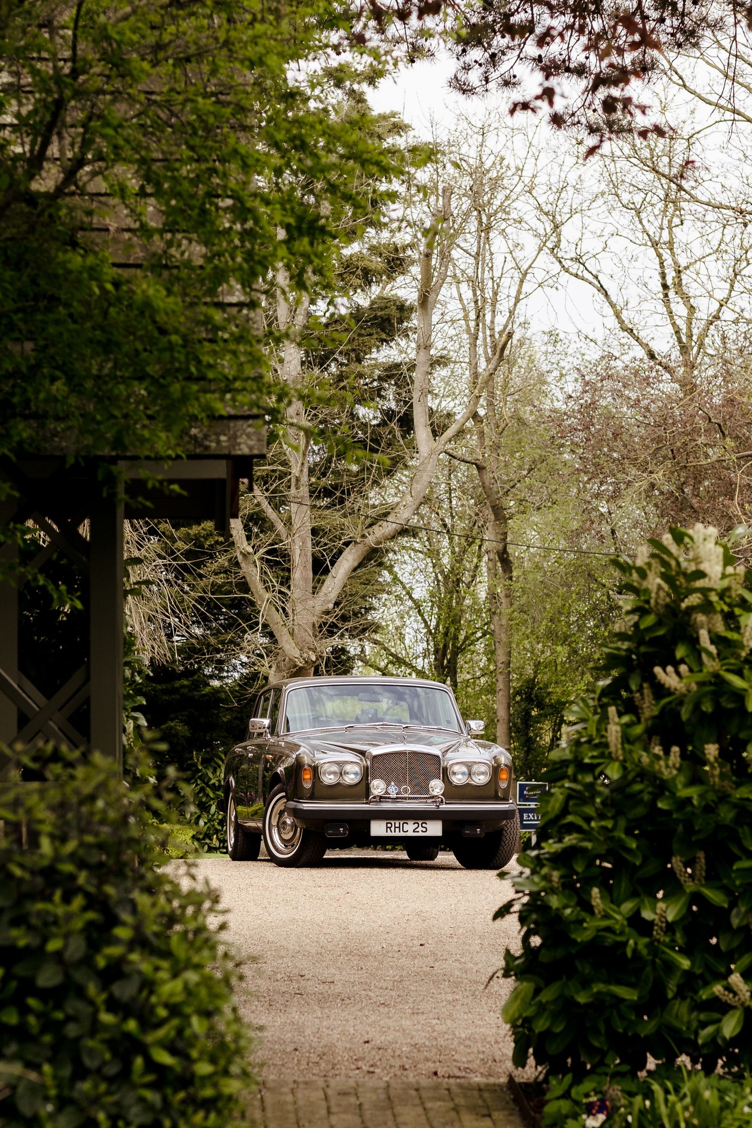 Vintage dark luxury car parked on a gravel driveway surrounded by lush green trees and hedges.
