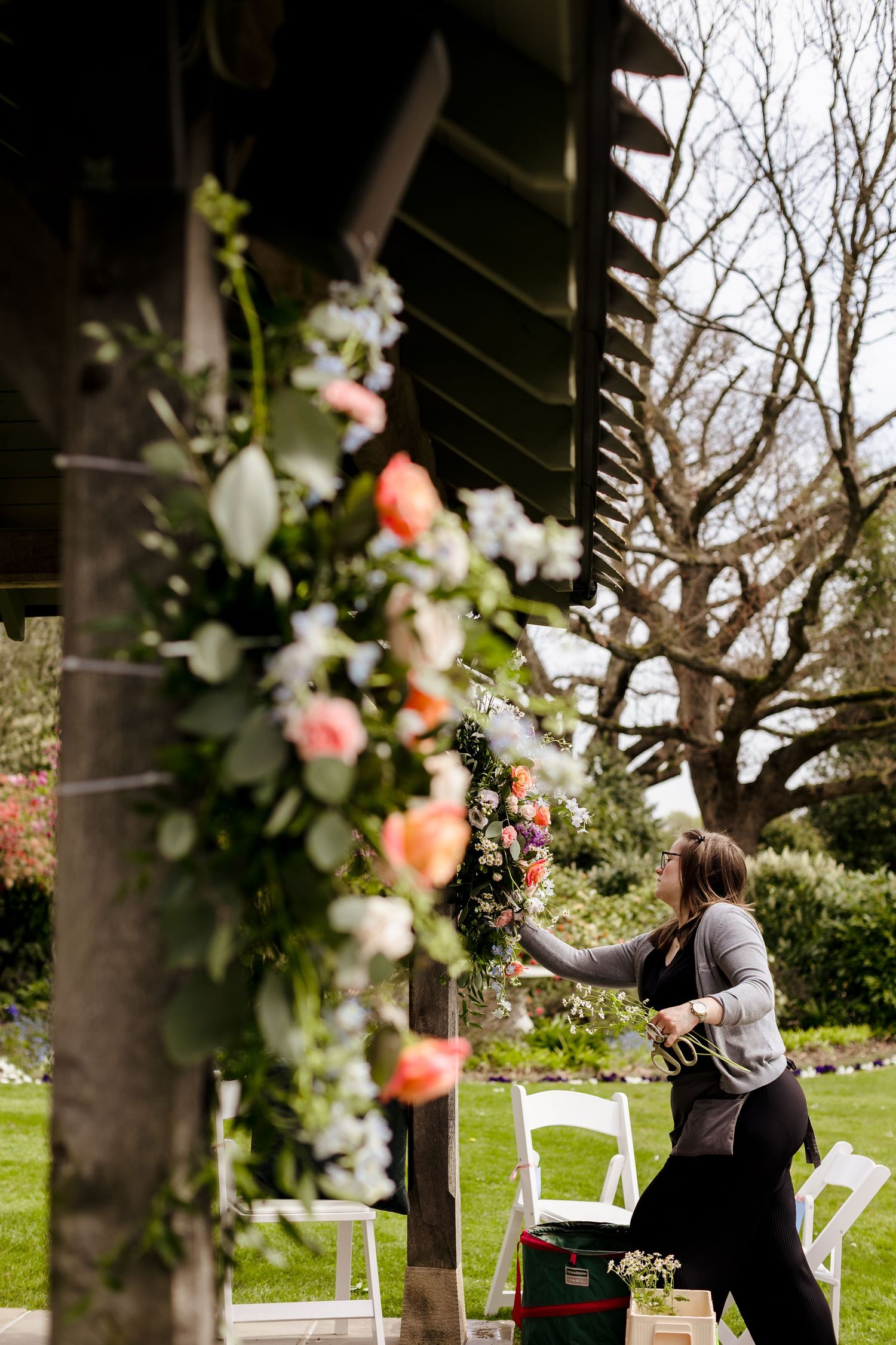 Florist arranging pink roses and greenery on a wooden pergola for an outdoor wedding ceremony.