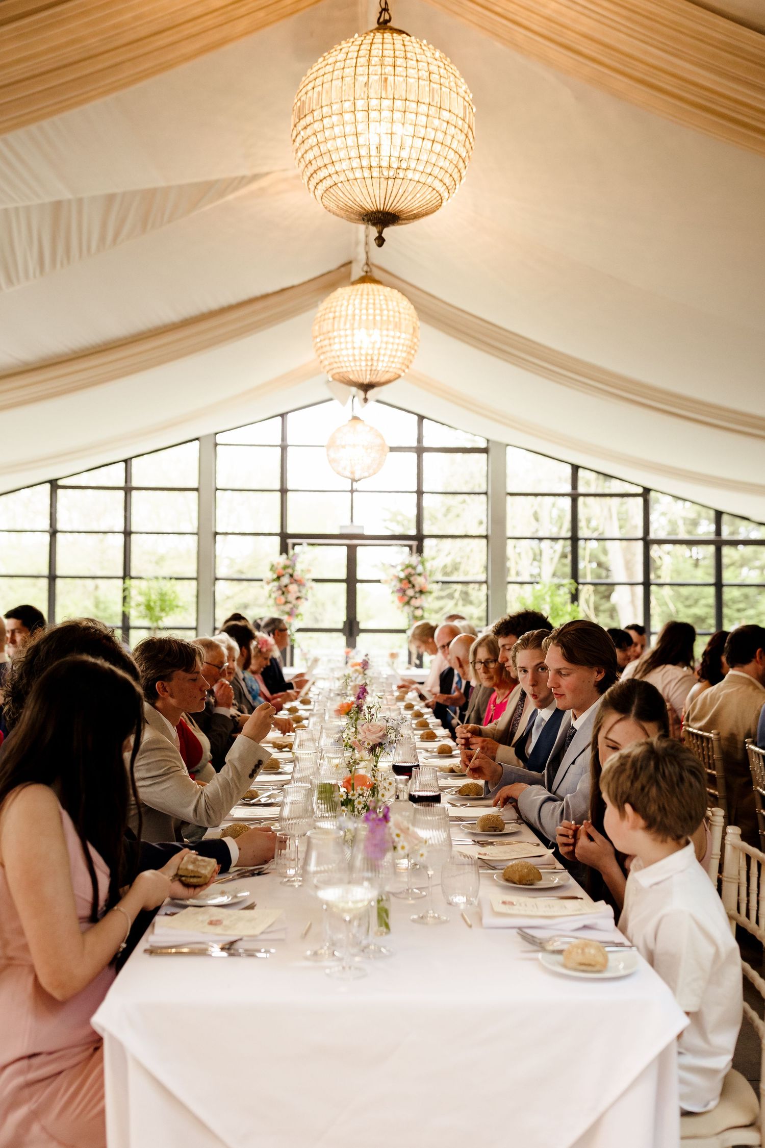 Wedding guests seated at long decorated tables in a marquee venue with globe chandeliers and large windows.