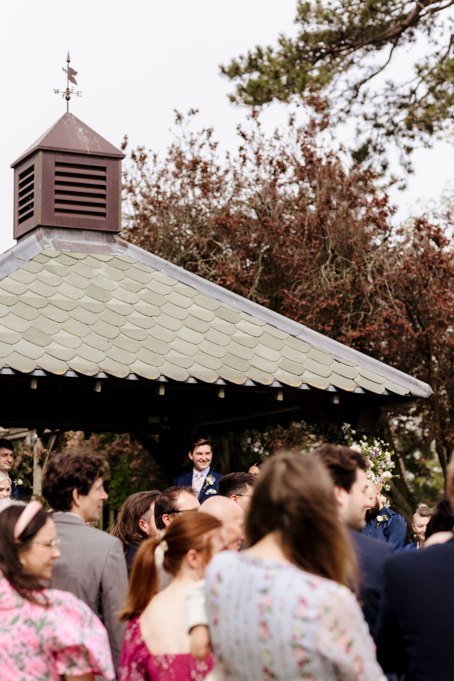 Groom waiting at outdoor wedding gazebo ceremony while guests gather on the lawn below.