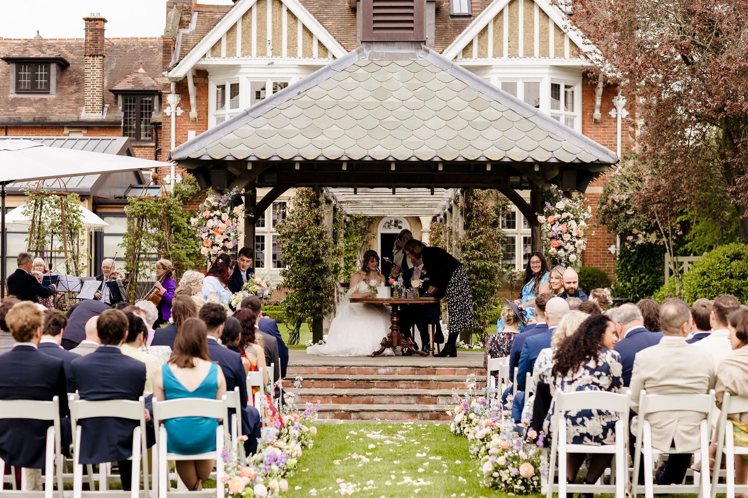 Outdoor wedding ceremony under ornate gazebo with floral arch and seated guests on lawn.