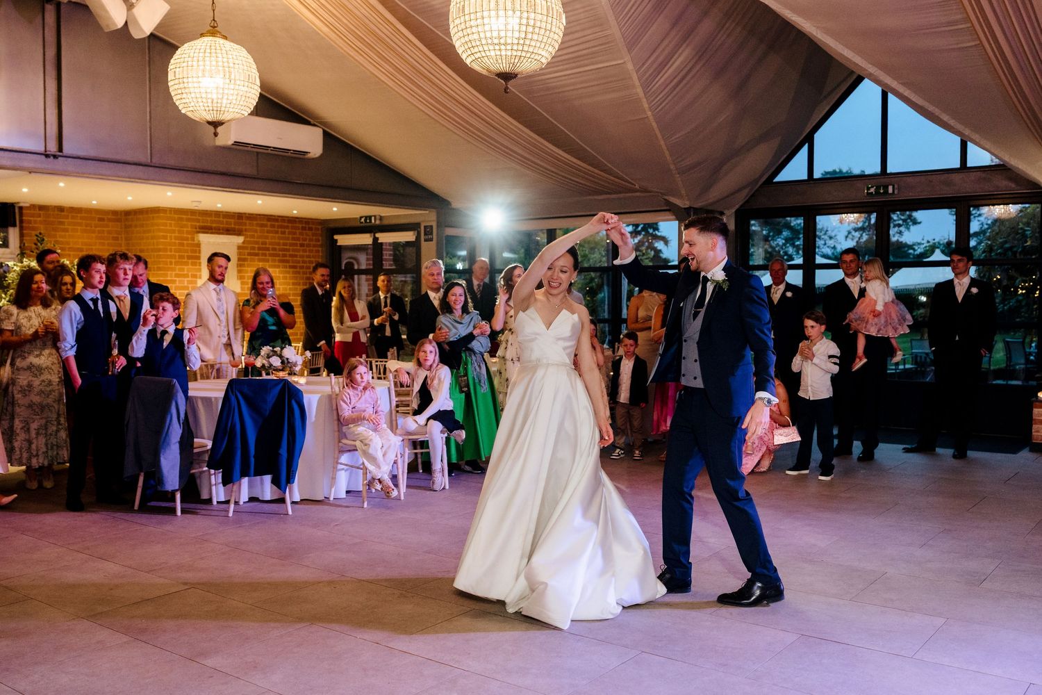 Bride in ballgown and groom in navy suit dance together at wedding reception with guests watching.
