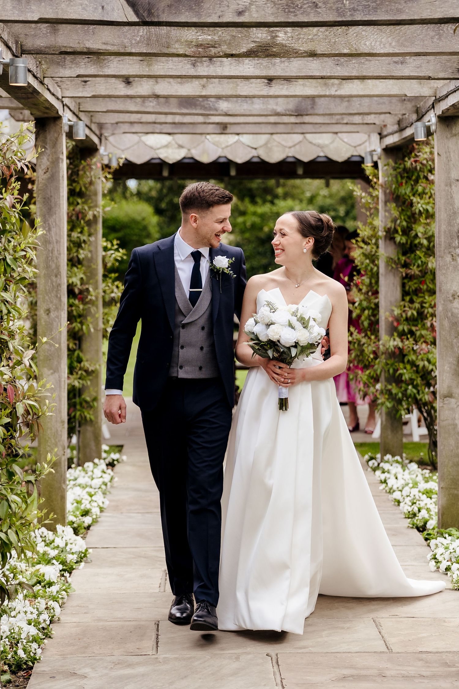 Bride and groom walking together under a floral pergola, bride holding a white bouquet on their wedding day.