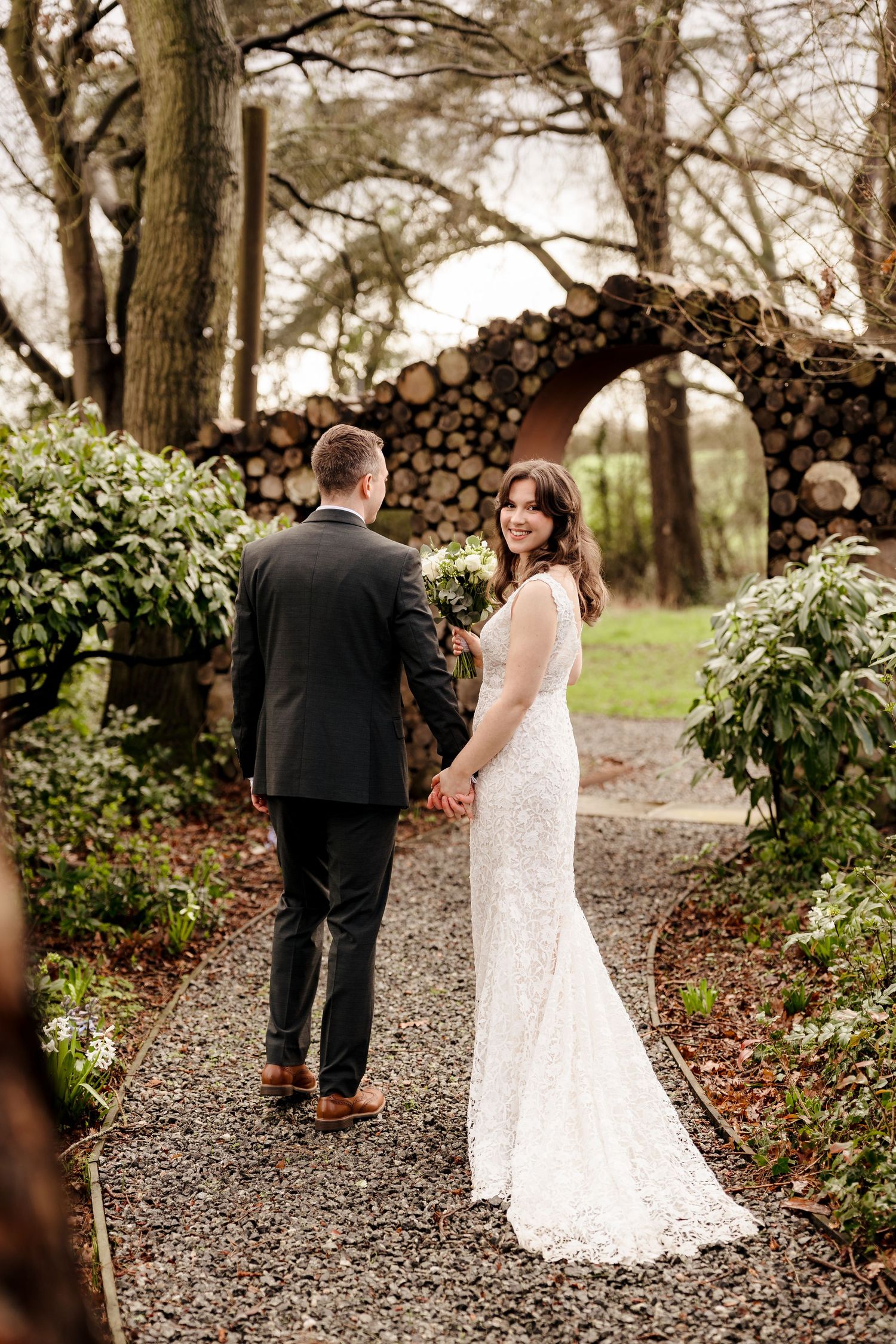 Bride in lace gown holds groom's hand on garden path near rustic log arch at wedding venue.