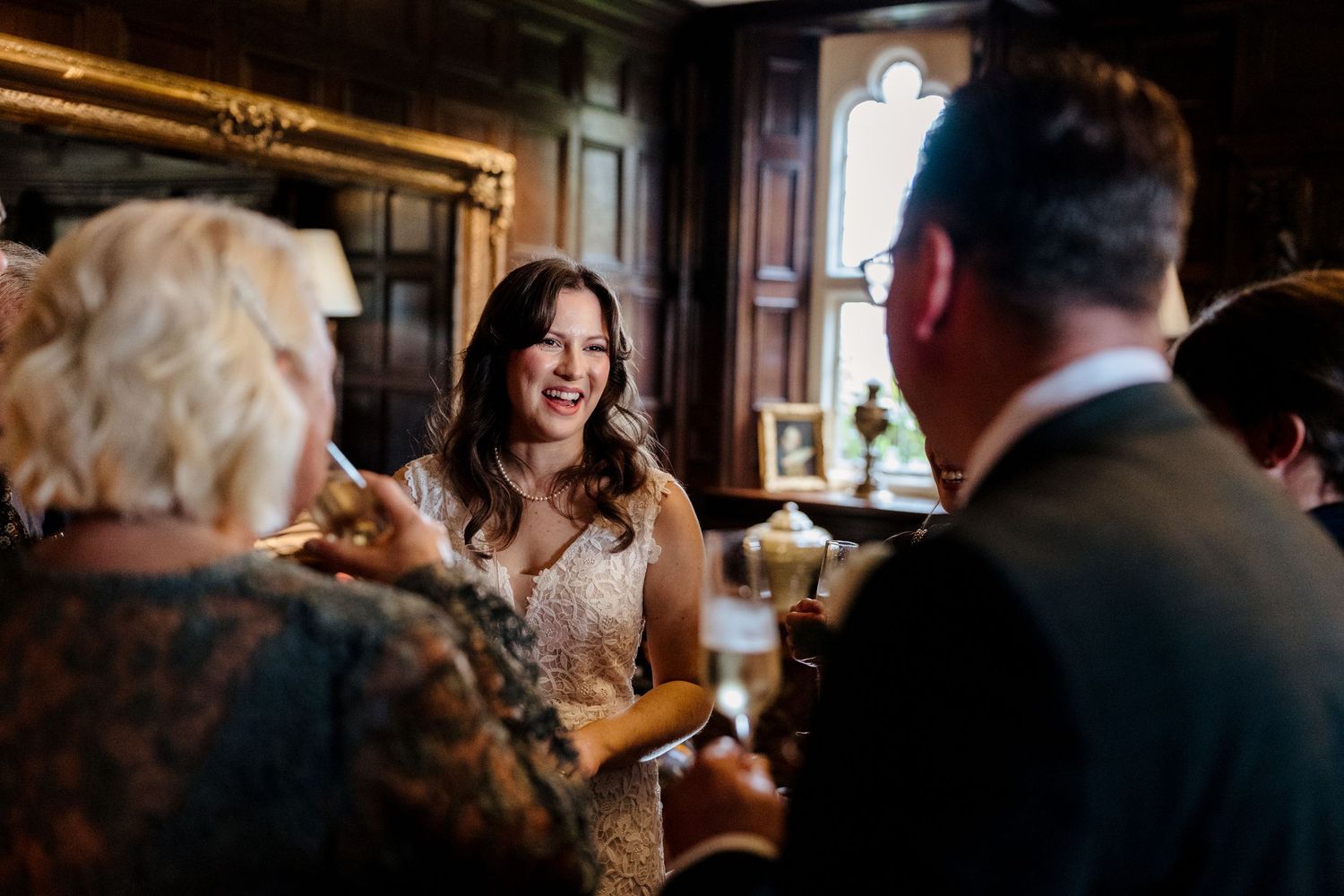 Radiant bride in lace gown laughs joyfully with guests during wedding reception in ornate room.