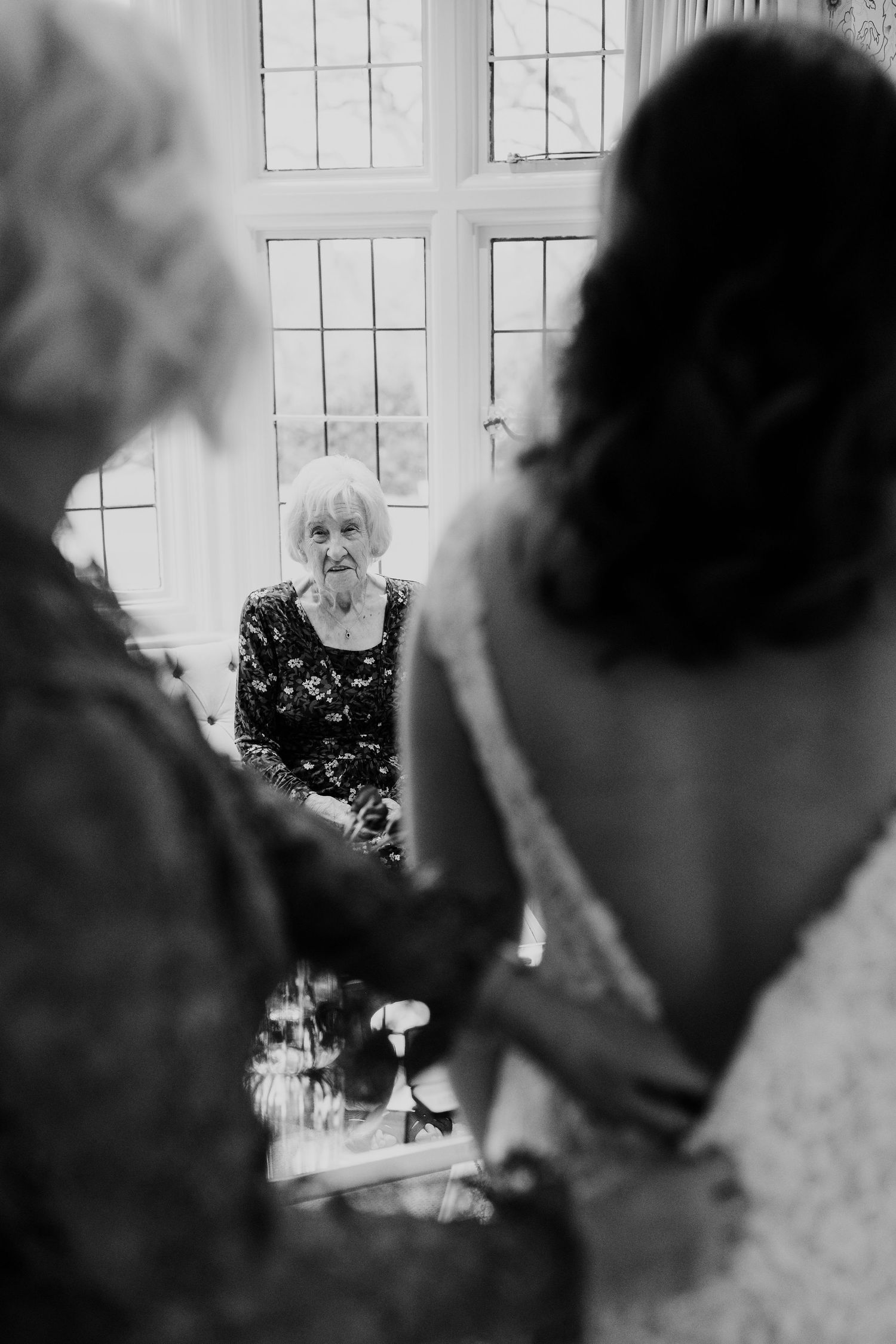 Black and white photo of elderly woman watching bride being helped into wedding dress by window.