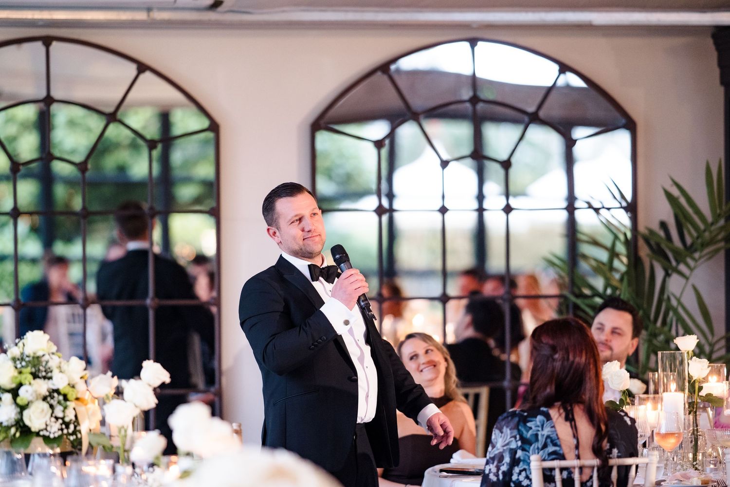 Groom in tuxedo delivering a heartfelt wedding speech at an elegant candlelit reception with floral decor.