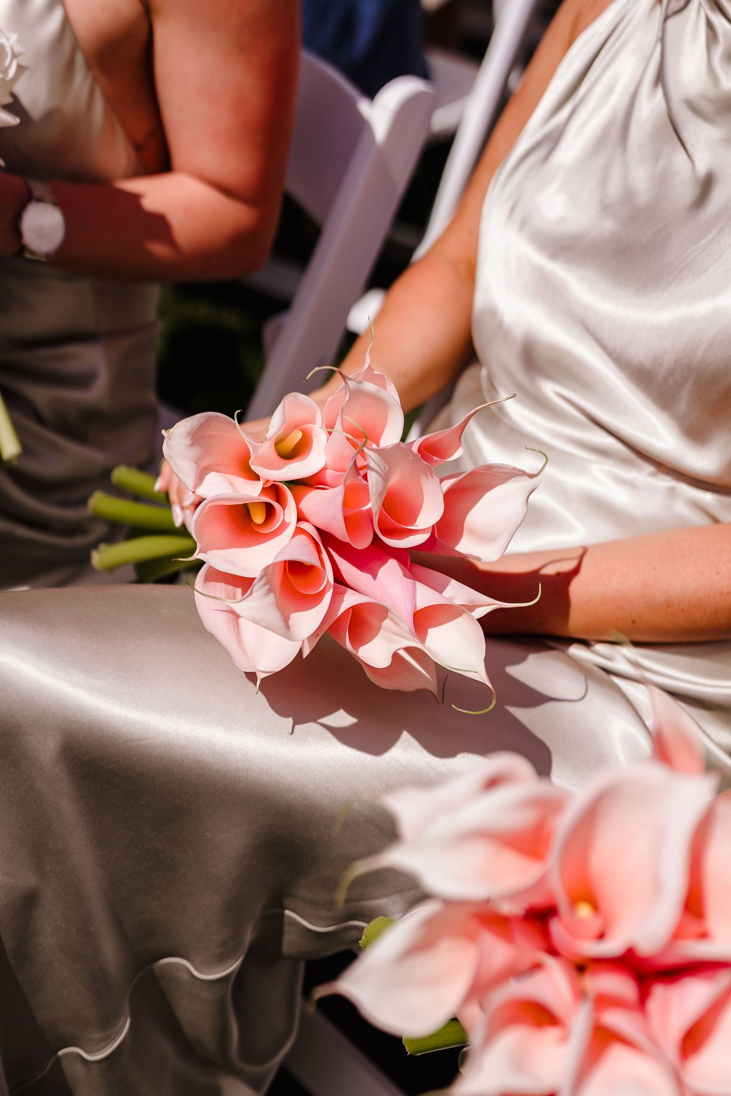 Bridesmaid in silver satin dress holds bouquet of pink calla lilies at outdoor wedding ceremony.