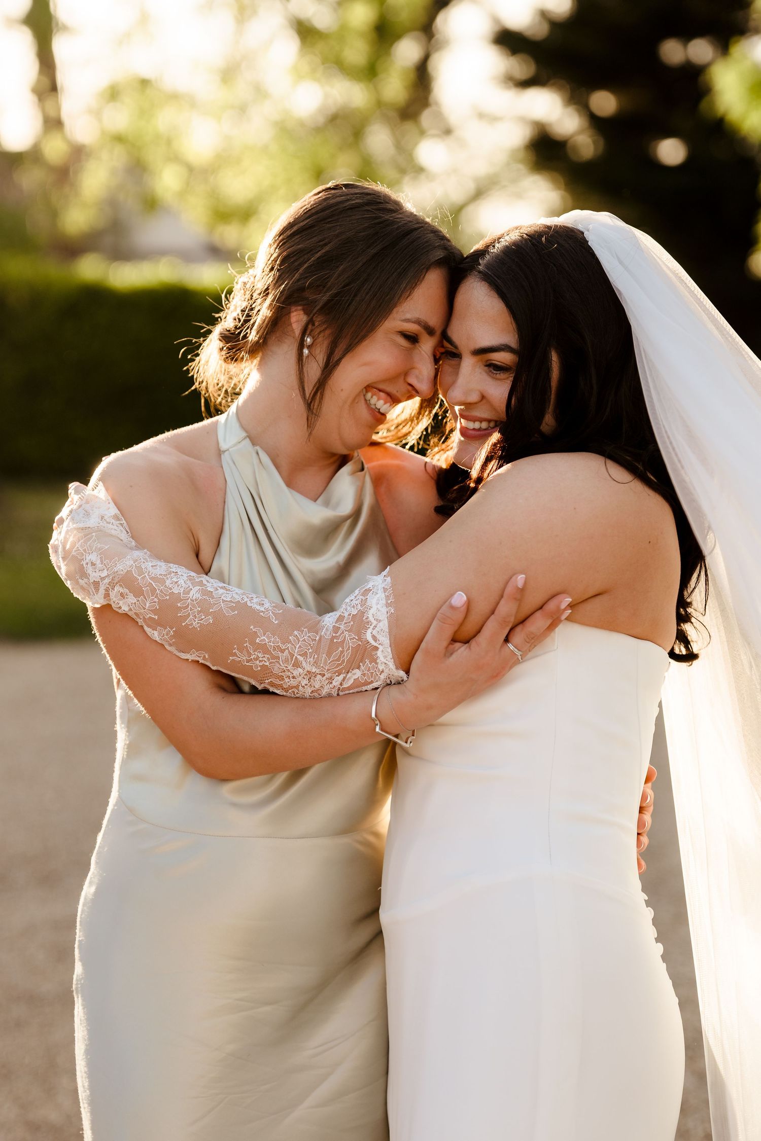 Bride in white gown and veil shares joyful embrace with bridesmaid in ivory dress in golden light.