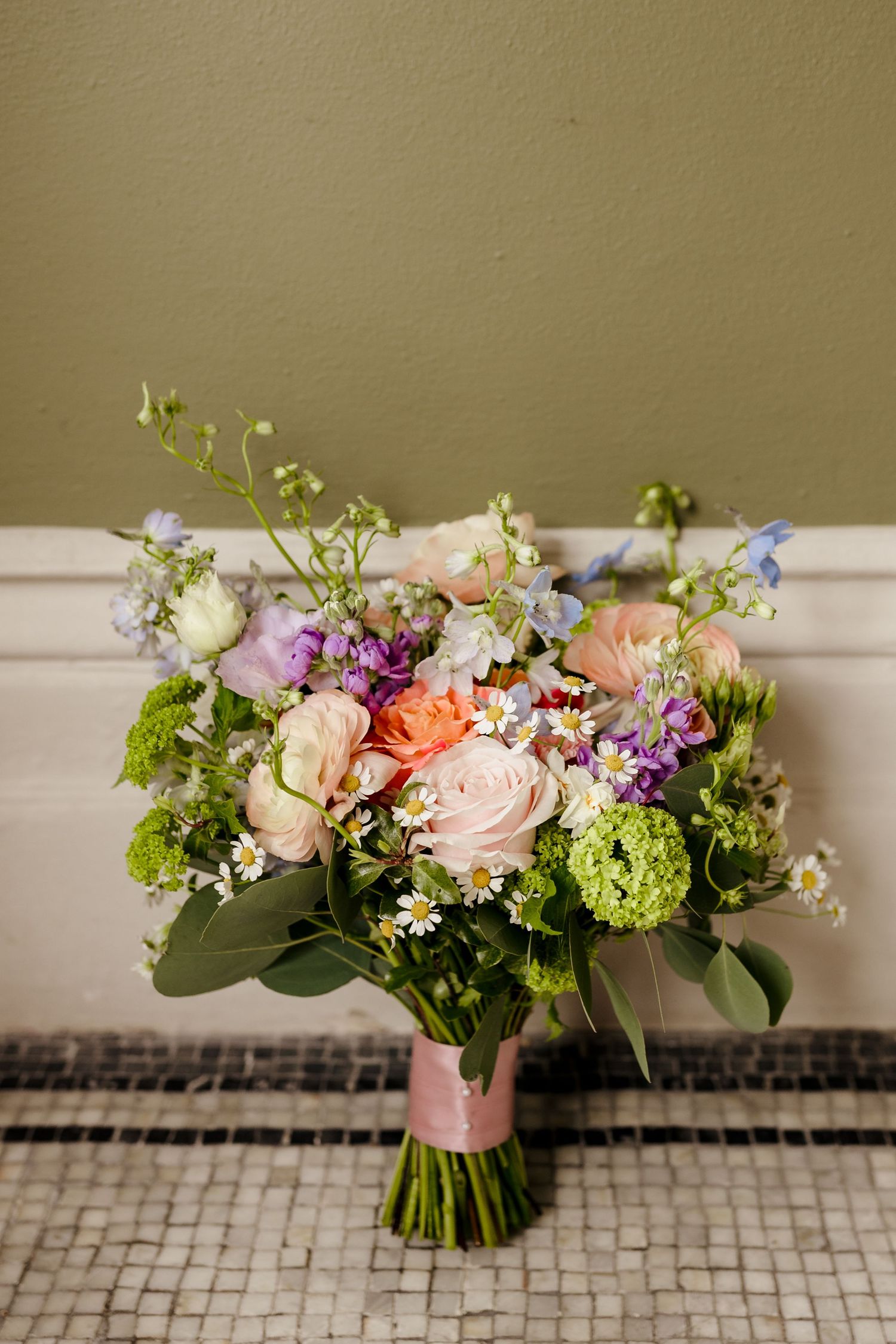 Colorful bridal bouquet featuring pink roses, purple flowers, green hydrangeas, and wildflowers tied with pink ribbon.