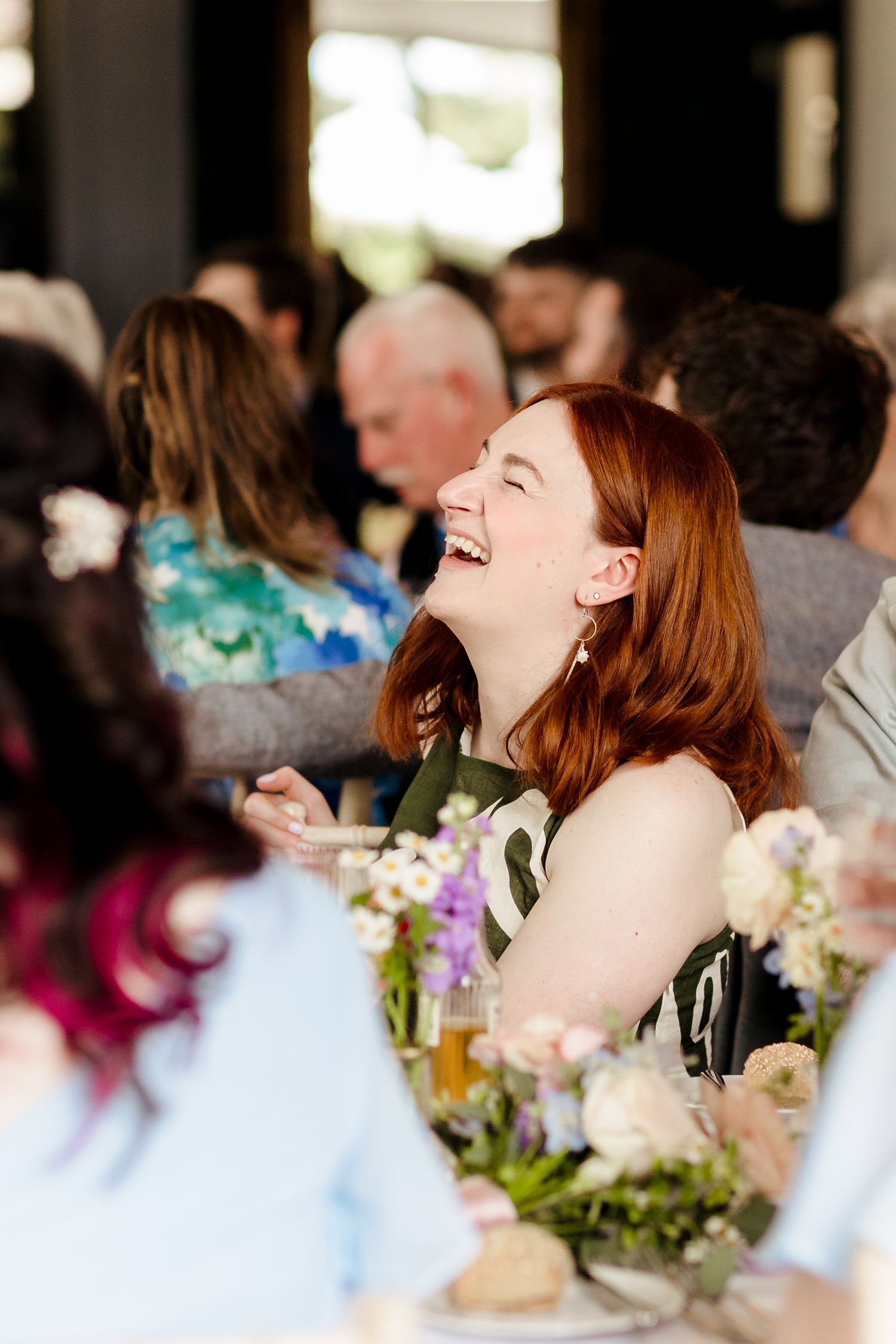 Joyful red-haired wedding guest laughing at reception table surrounded by floral centerpieces and guests.