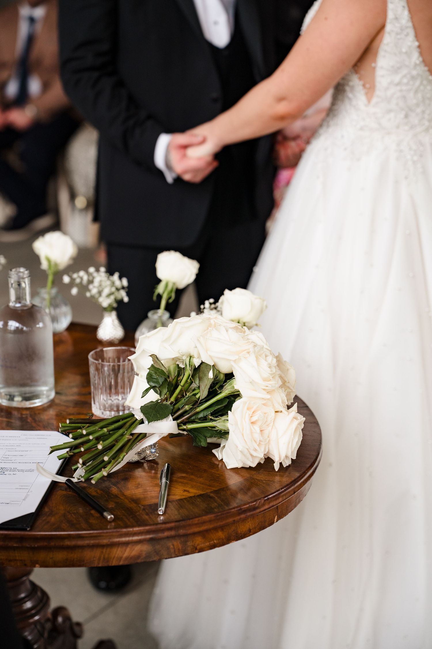 White rose bridal bouquet resting on a wooden table during a wedding ceremony with couple holding hands.