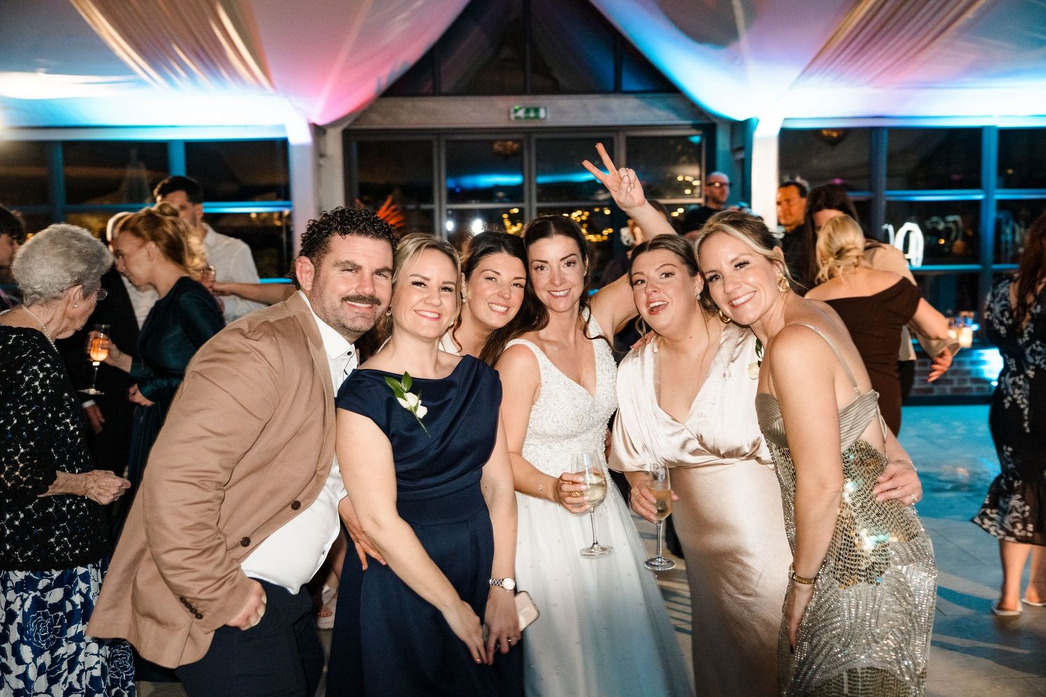 Group of smiling wedding guests posing together at a festive evening reception with blue ambient lighting.