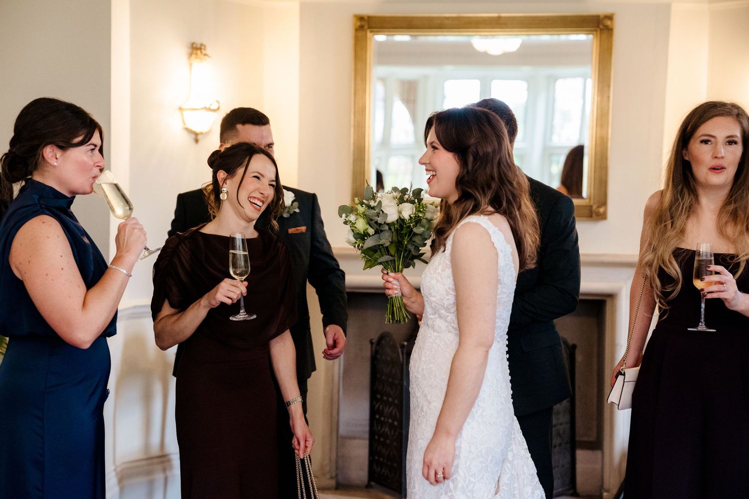 Bride in lace dress laughs with bridesmaids holding champagne glasses and bouquet during wedding reception.